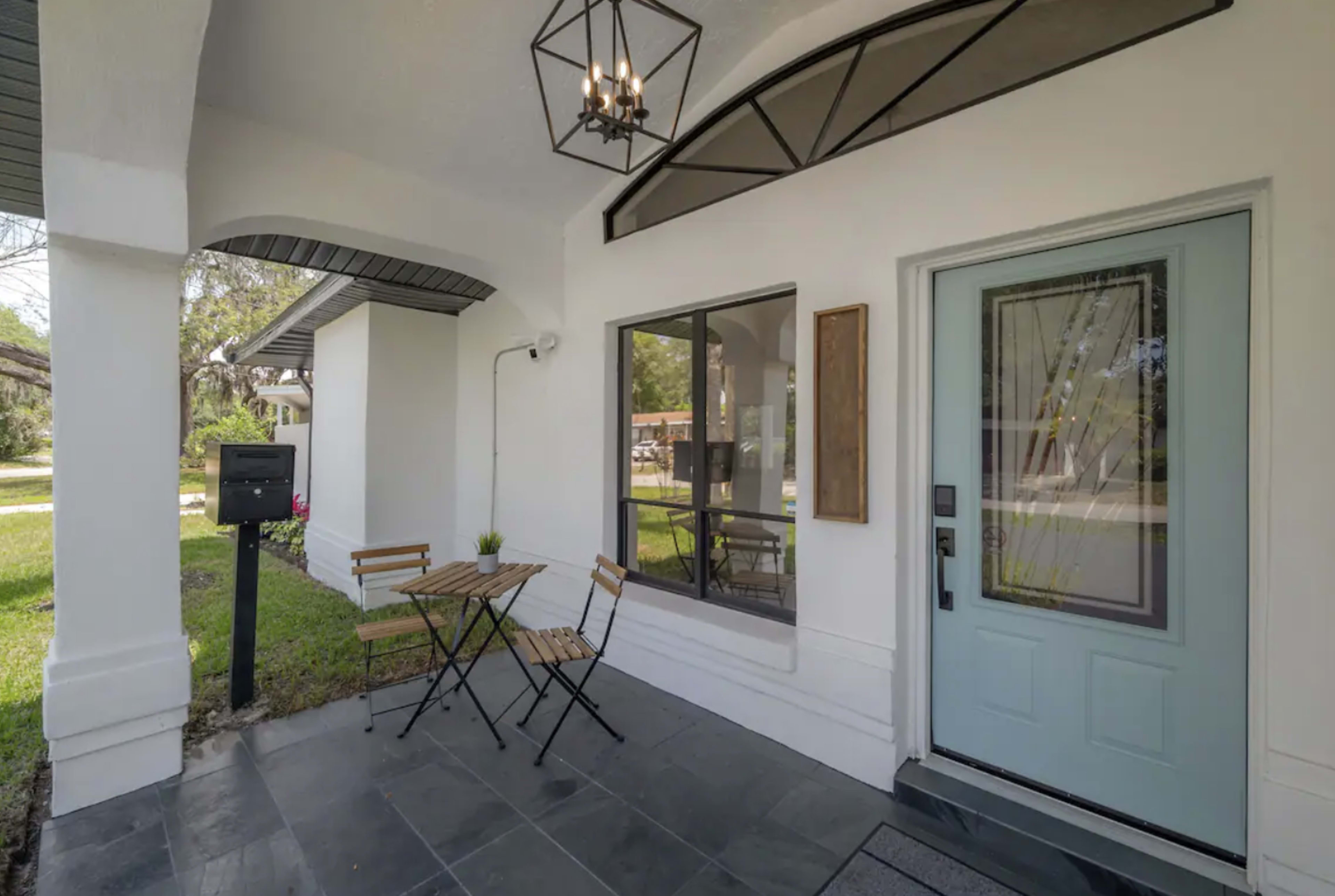 The image shows a small front porch with a blue door, a light fixture above, and a bistro table with two chairs.