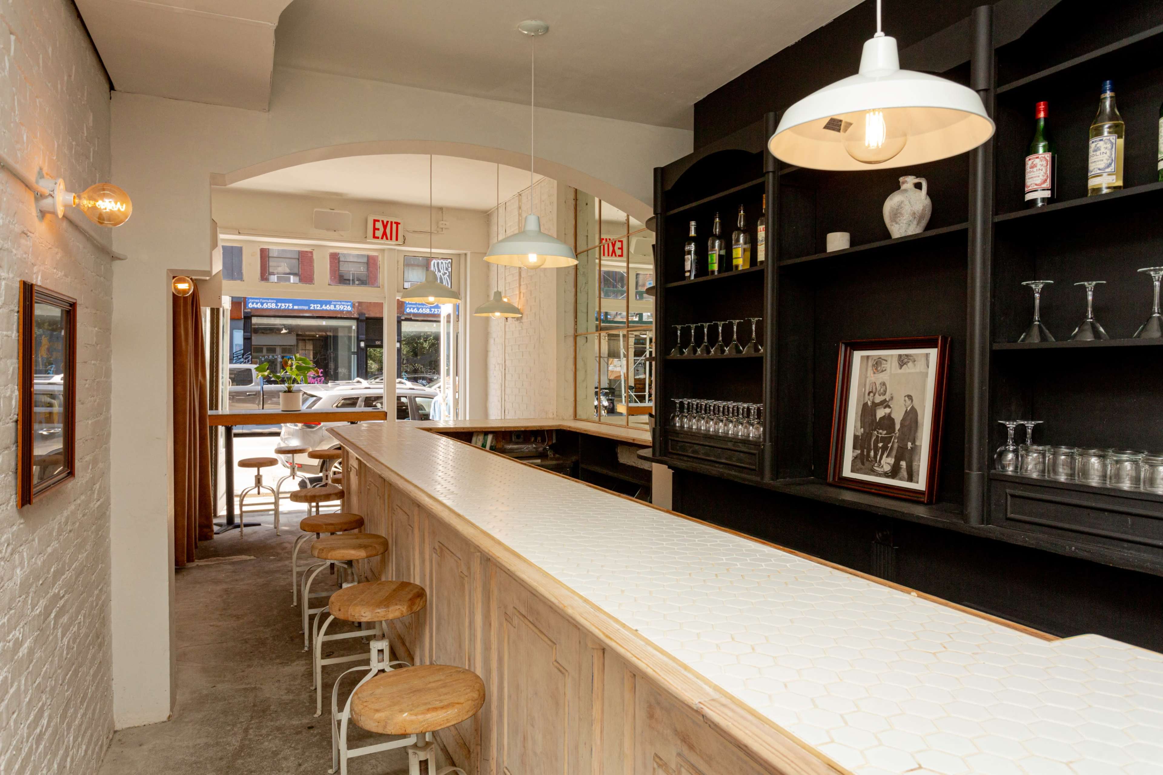 The image shows a bar area with a long, hexagon-tiled counter and several wooden stools, framed by white walls and a window displaying the street outside.
