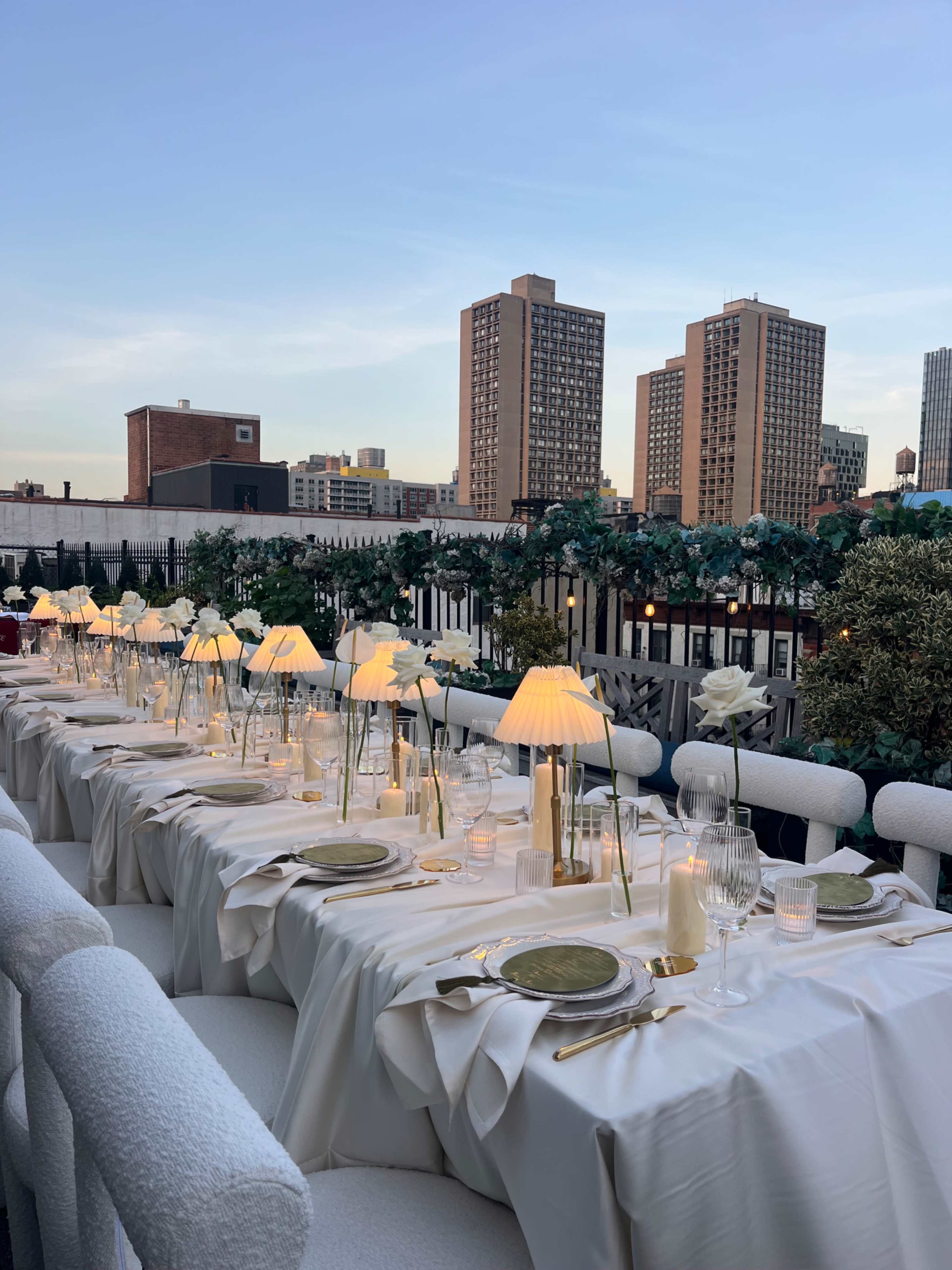 A long dining table adorned with elegant tableware and lamps is set on a rooftop overlooking city buildings at sunset.