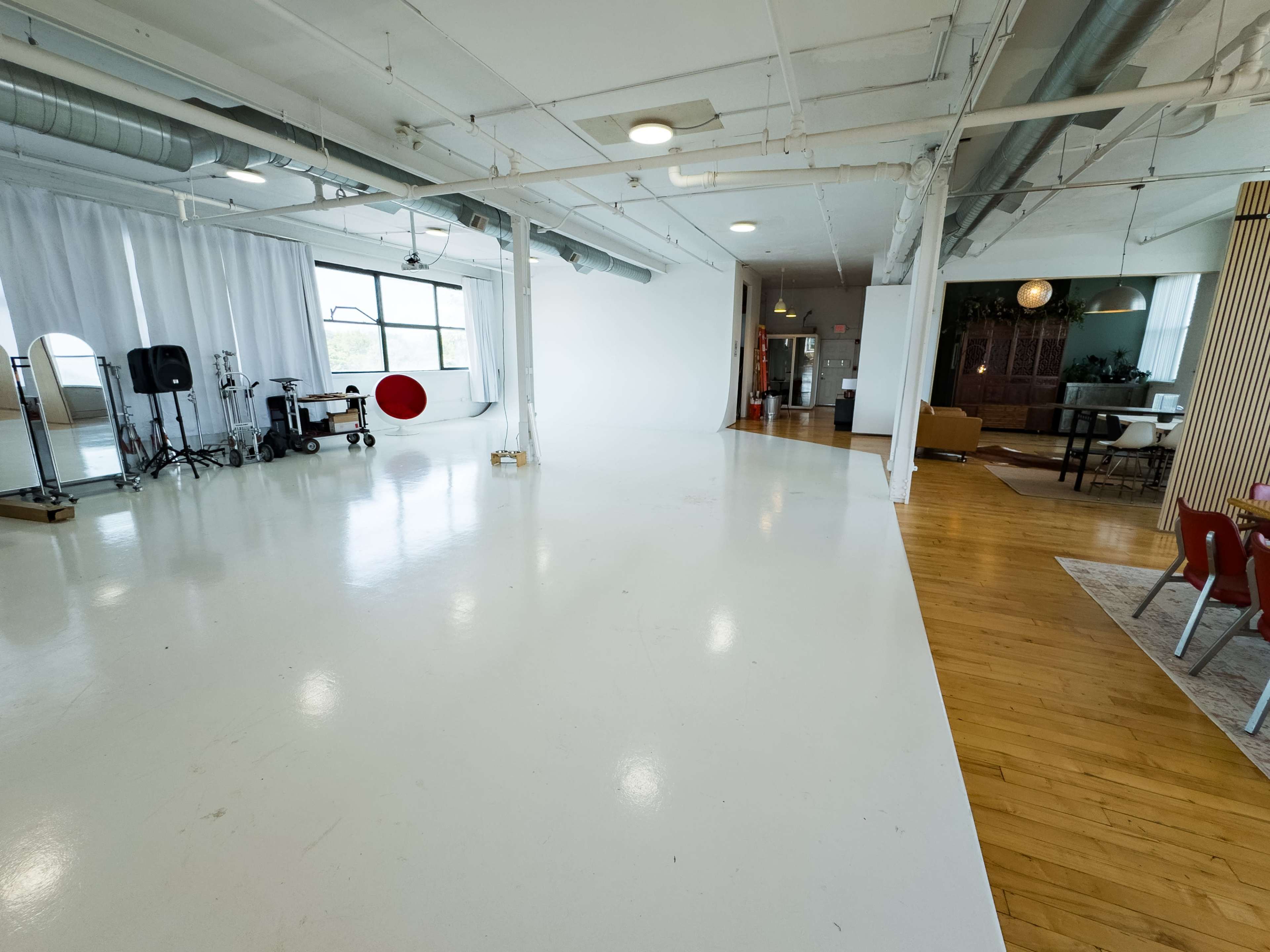 The image shows a spacious, well-lit studio with white floors, large windows, and a mix of furniture and equipment in two separate areas.