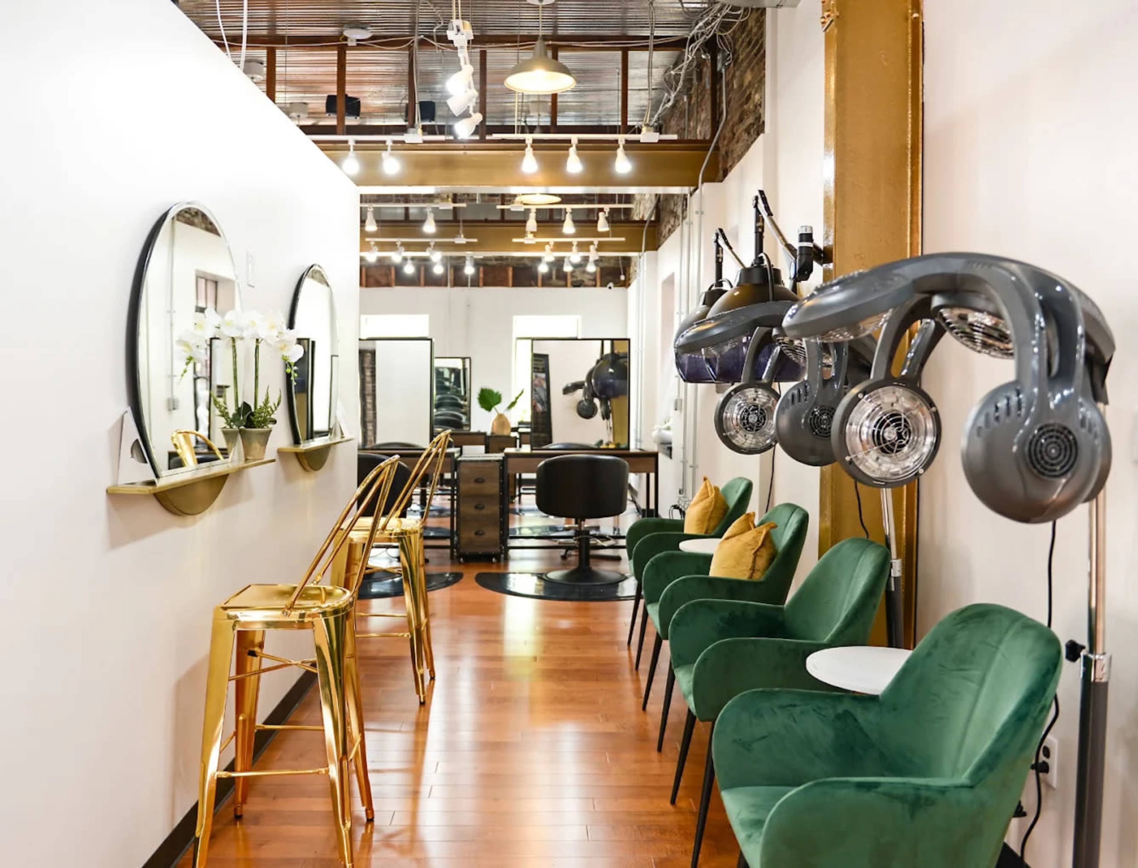 The image shows a modern hair salon with green chairs, gold accents, and large mirrors along the walls, featuring hair dryers mounted on stands.