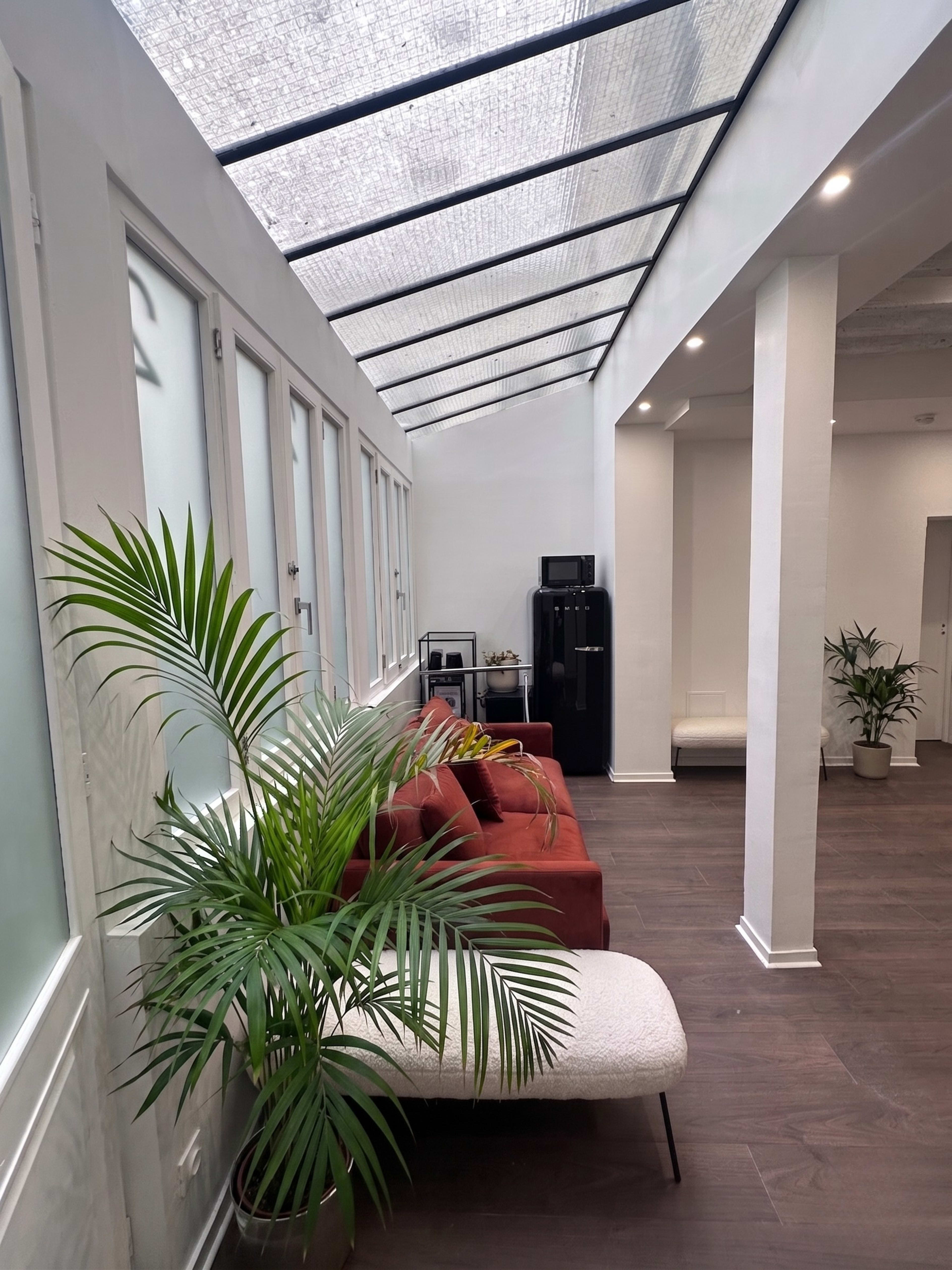 The image shows a bright interior space featuring a long, narrow room with large windows, a red sofa, and potted plants.