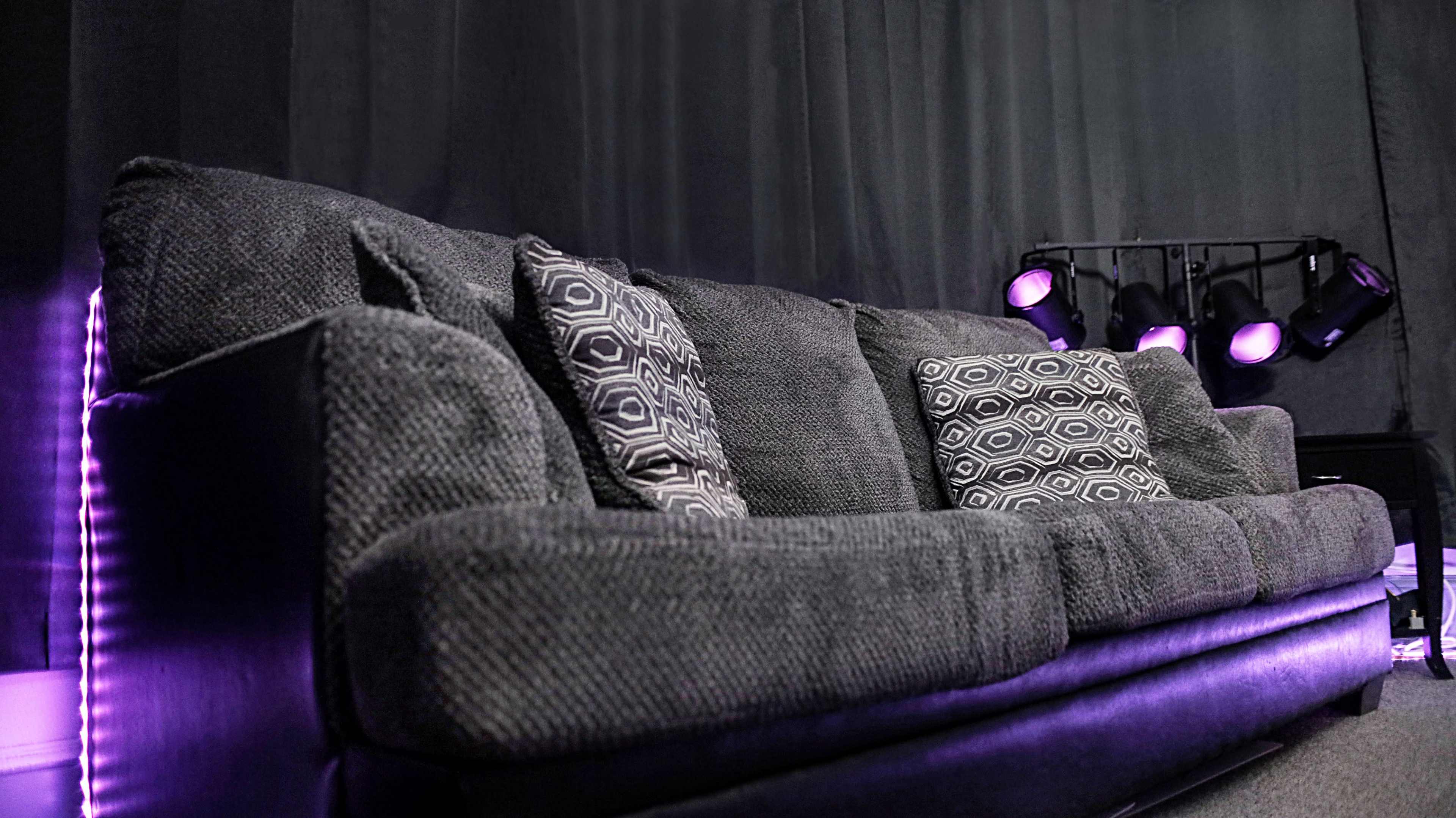 A gray couch with decorative pillows is positioned against a dark backdrop, illuminated by stage lights in the background.