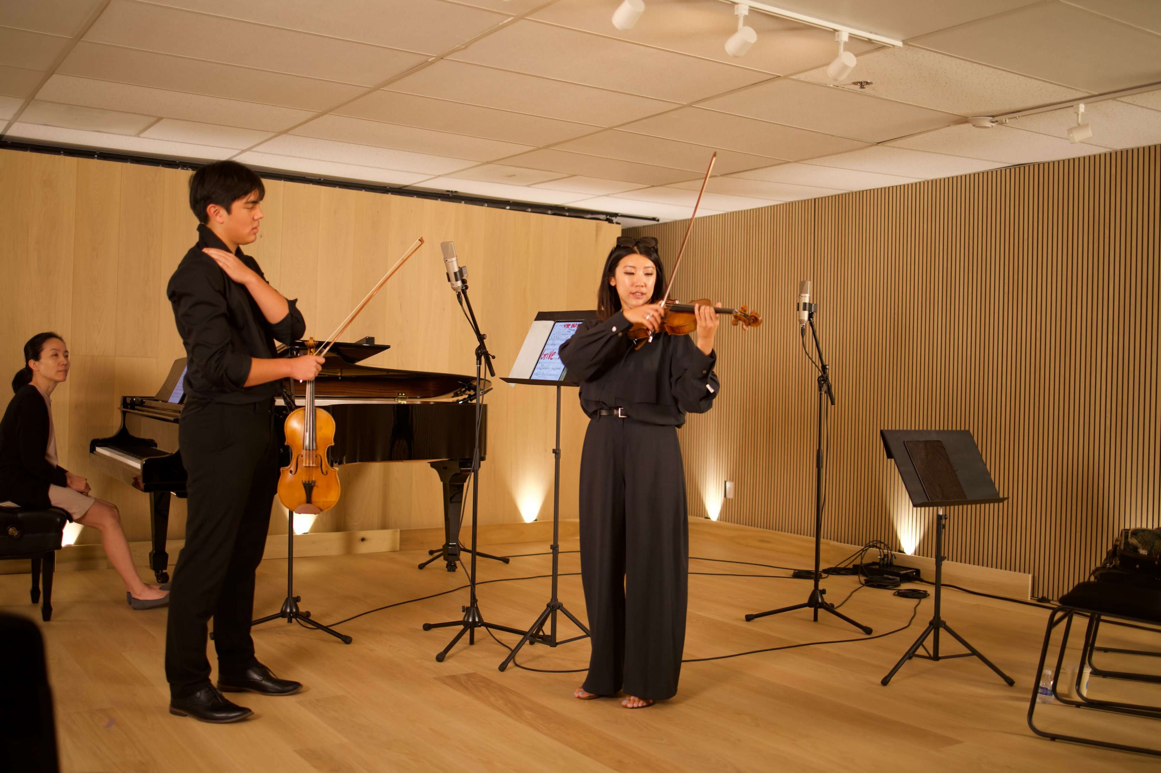 A violinist performs in front of an audience in a modern concert space, accompanied by a pianist.
