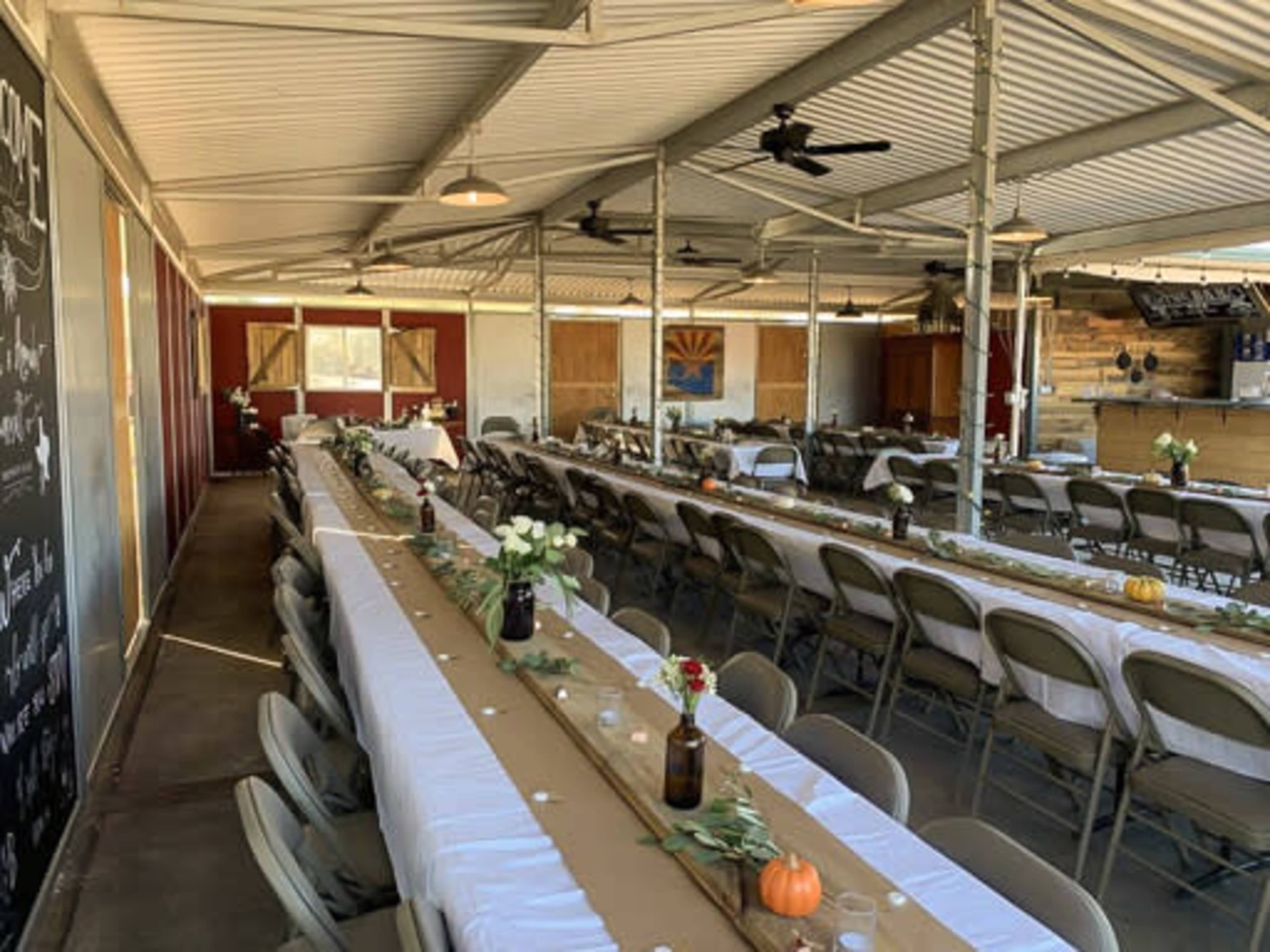 A spacious dining area with long tables arranged in rows, adorned with white tablecloths and simple centerpieces, under a metal roof with visible fans.