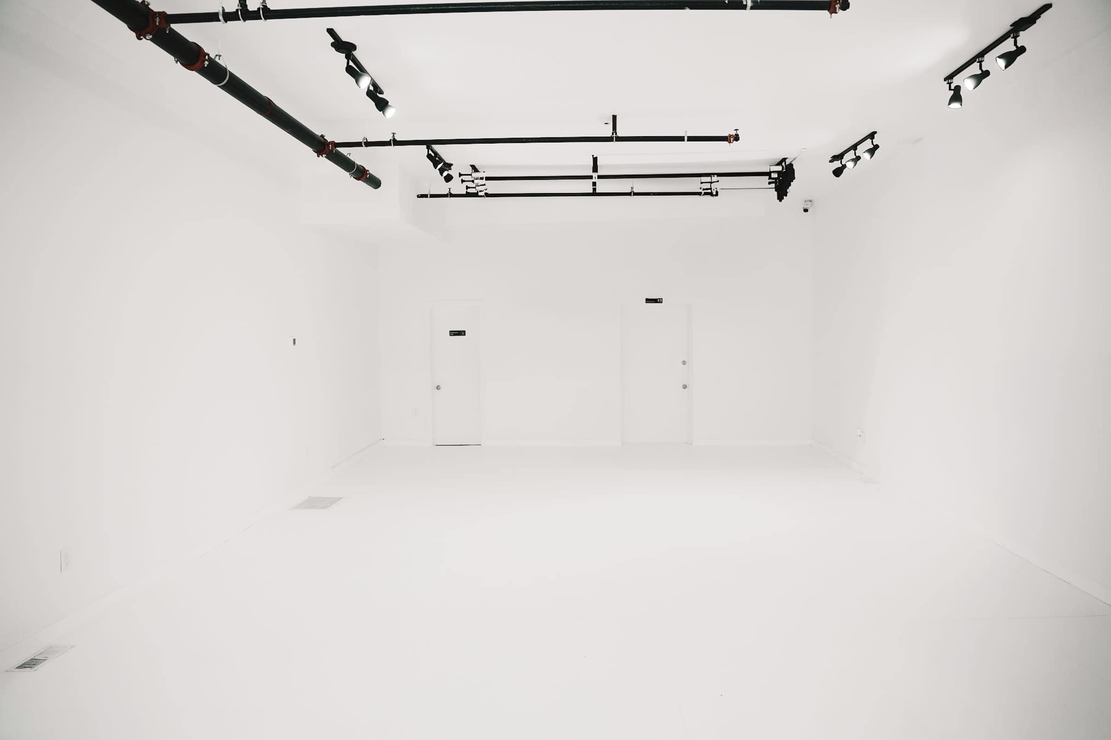 The image shows a large, empty white room with two doors and overhead lighting fixtures along the ceiling.
