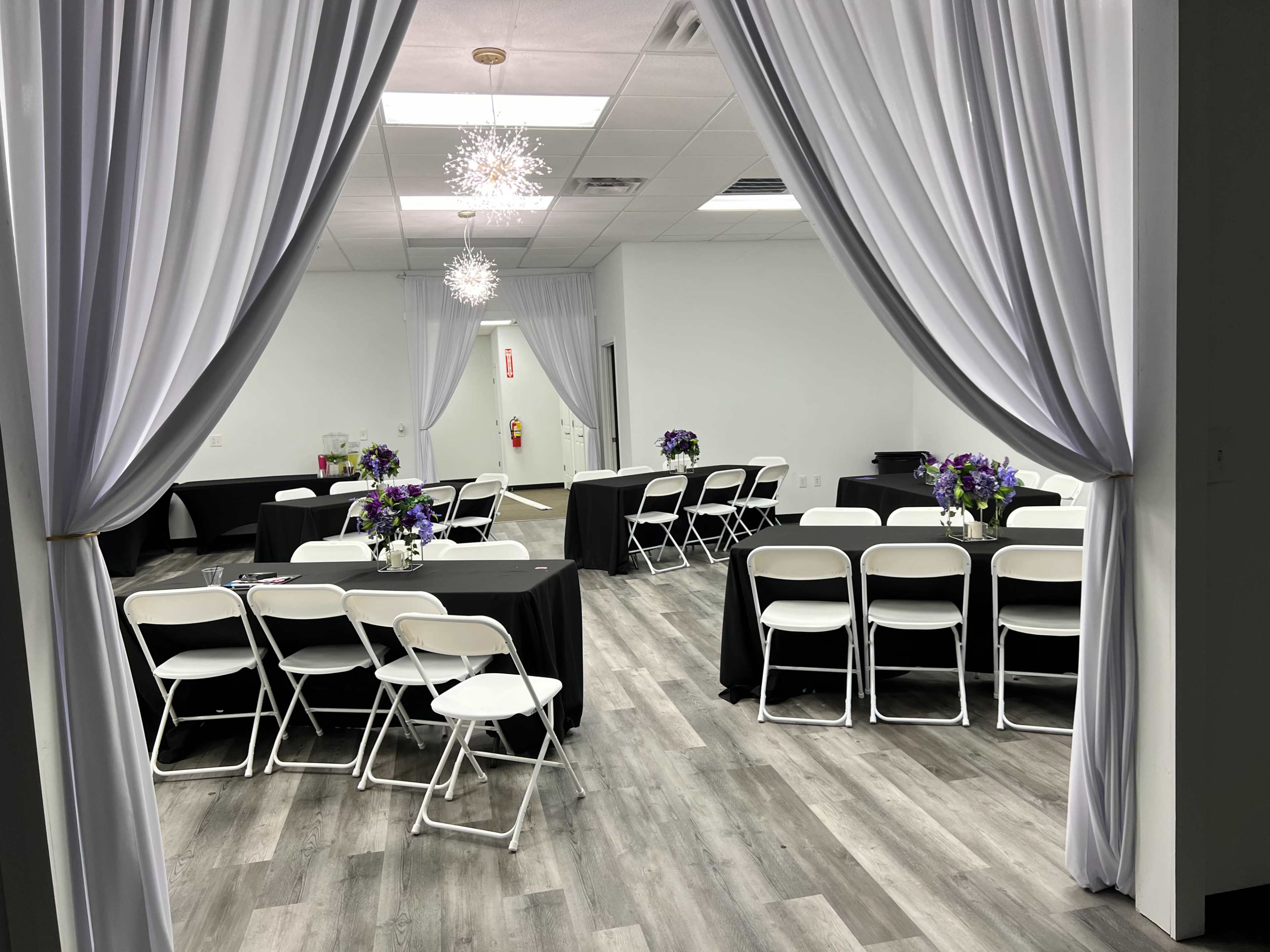 The image shows a room set up for an event, featuring round tables with black tablecloths, white folding chairs, and floral centerpieces.