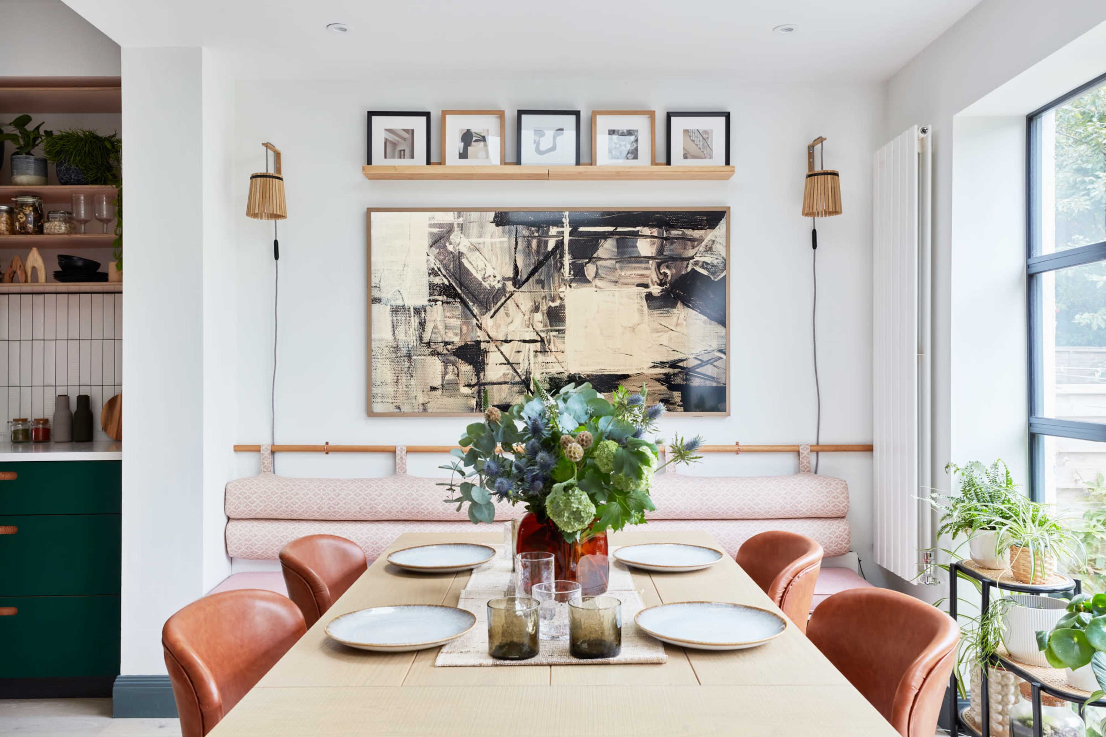 A dining area features a wooden table set for six, surrounded by leather chairs, with a large abstract painting and framed photos displayed on the wall above a cushioned bench.
