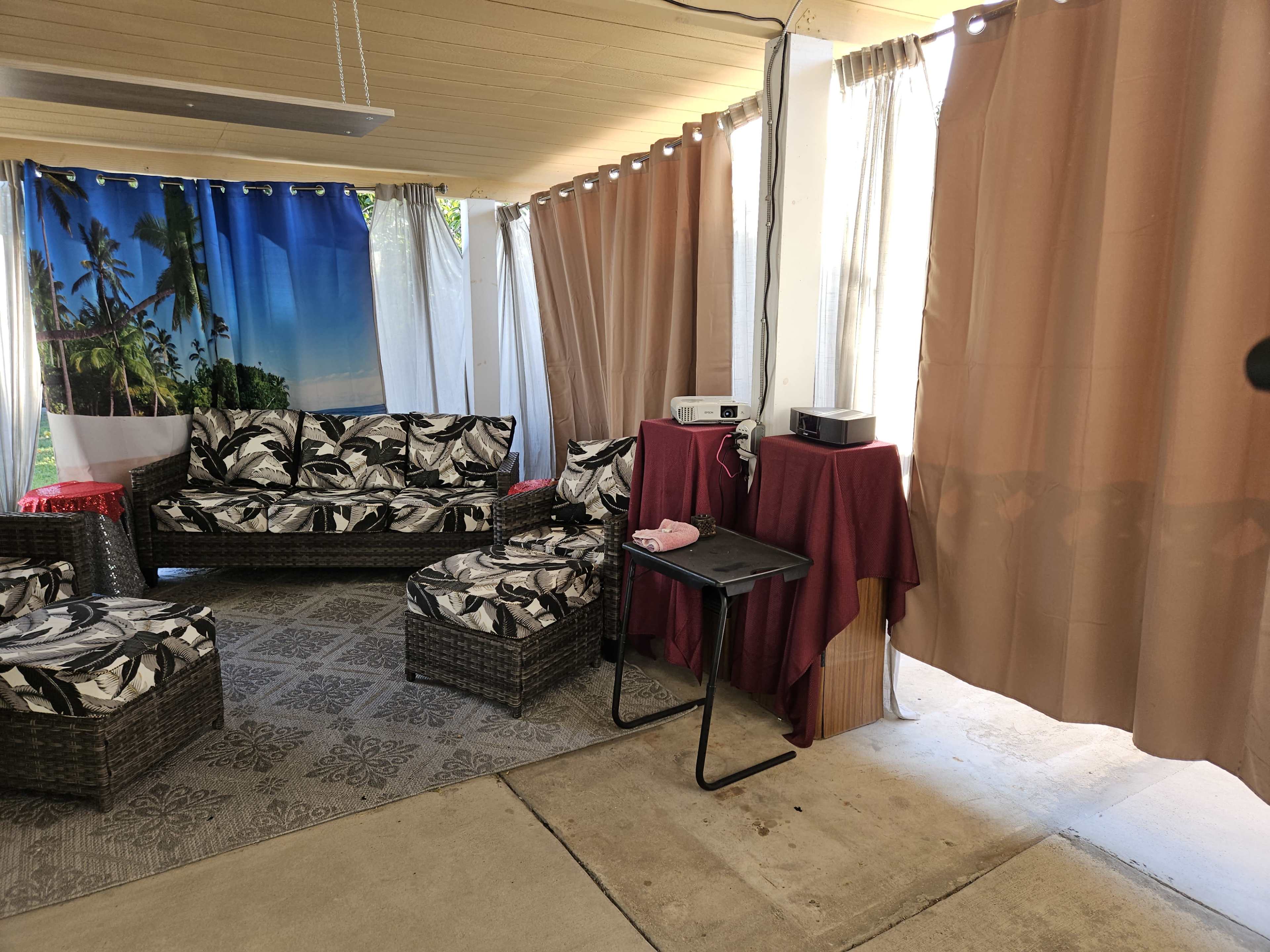 A covered outdoor seating area with wicker furniture, a projector setup, and curtains separating the space.