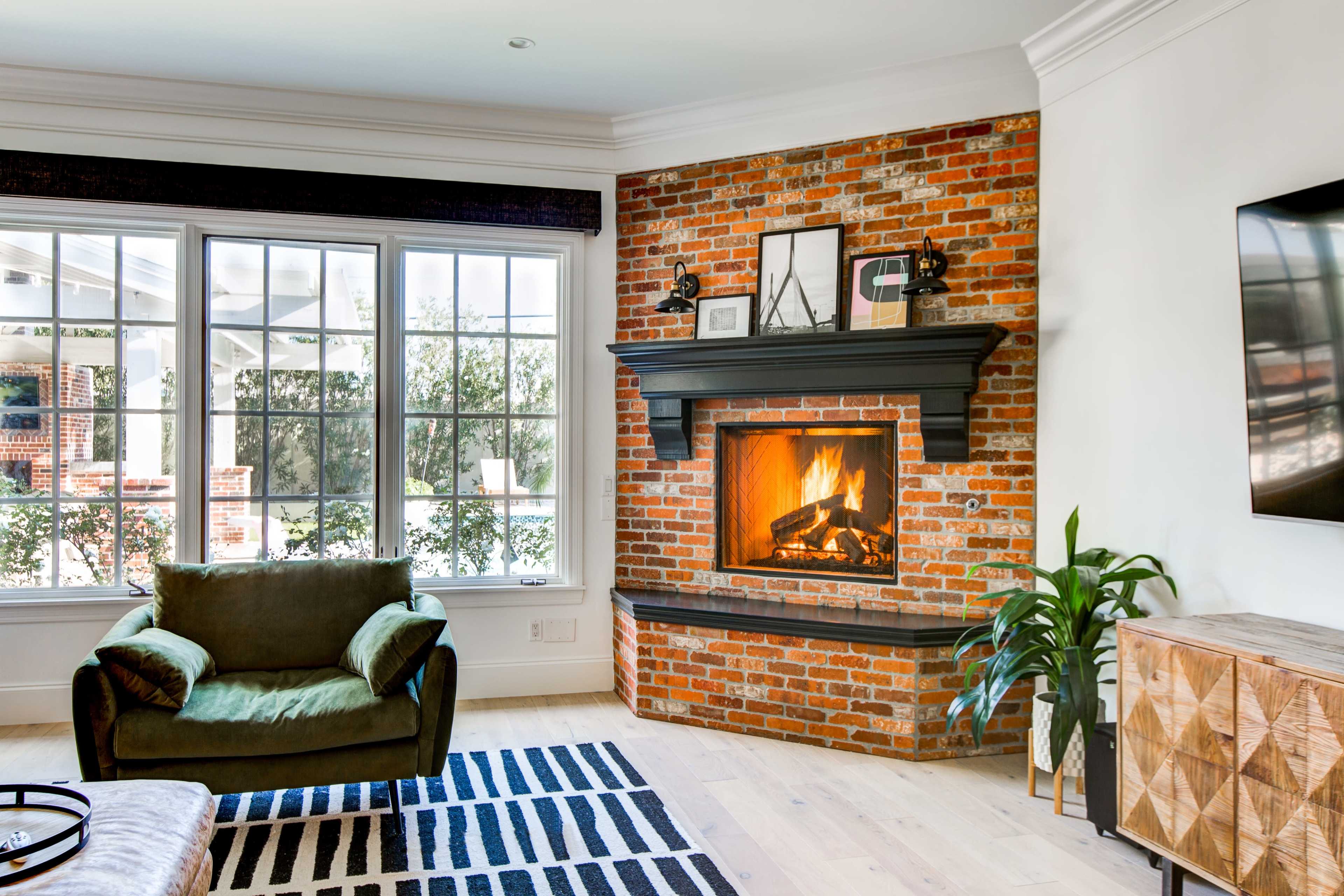 A living room with a brick fireplace, a green armchair, large windows, and a patterned rug.