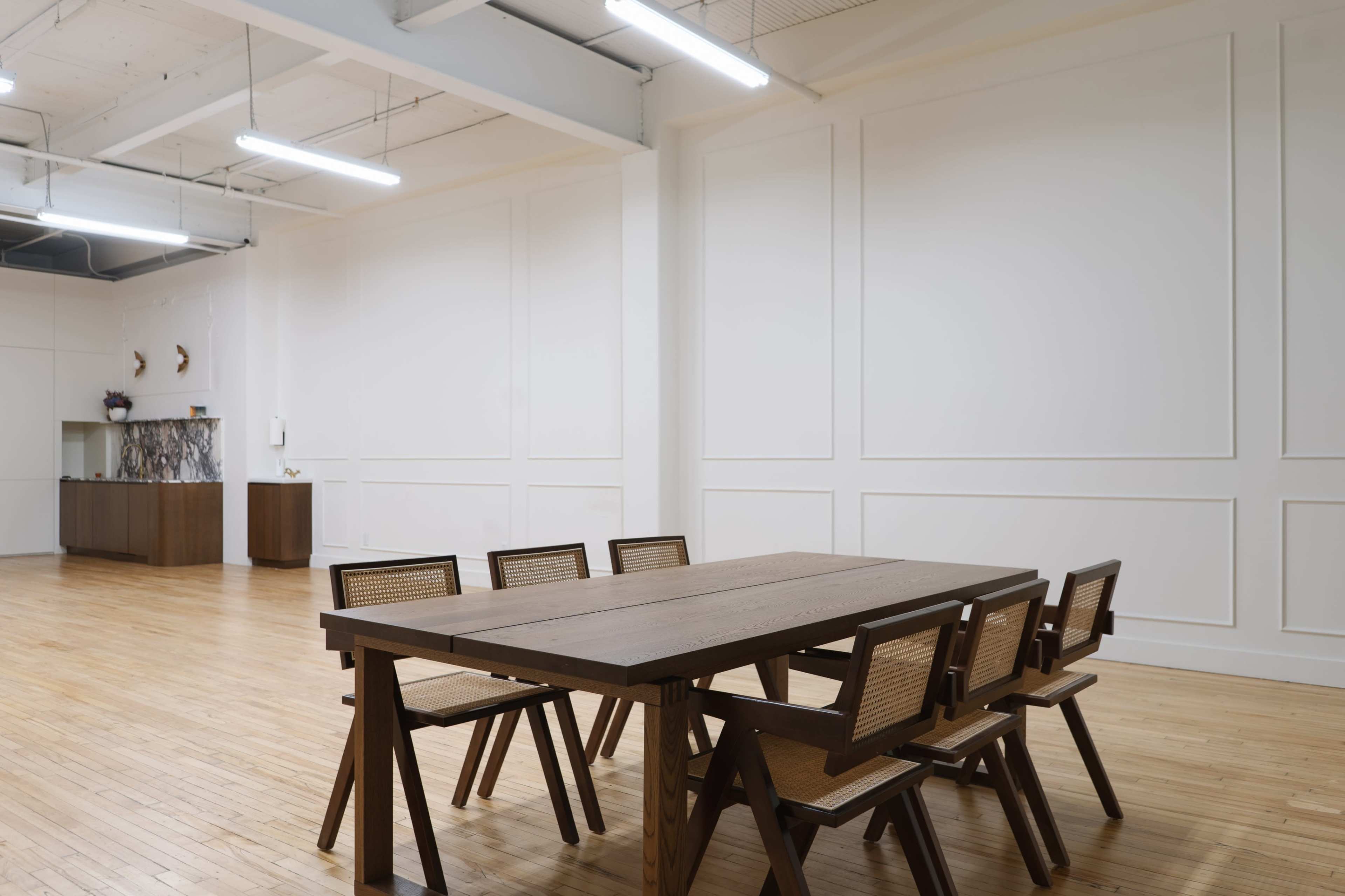 A spacious room features a large wooden table surrounded by six chairs, with plain white walls and a minimalist design.