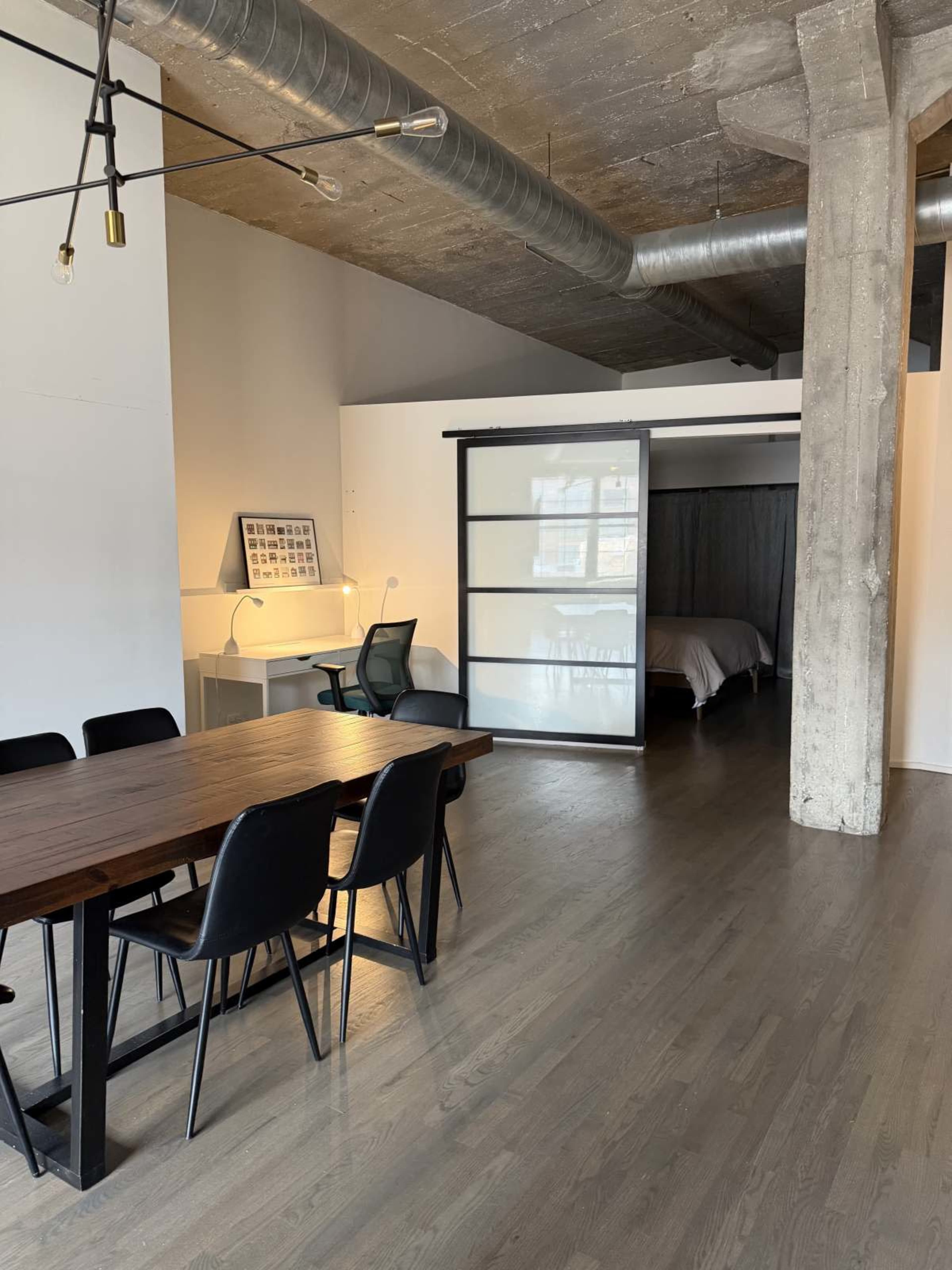 The image shows a modern interior space featuring a dining table with chairs, a small desk area, and a sliding door that leads to a bedroom.
