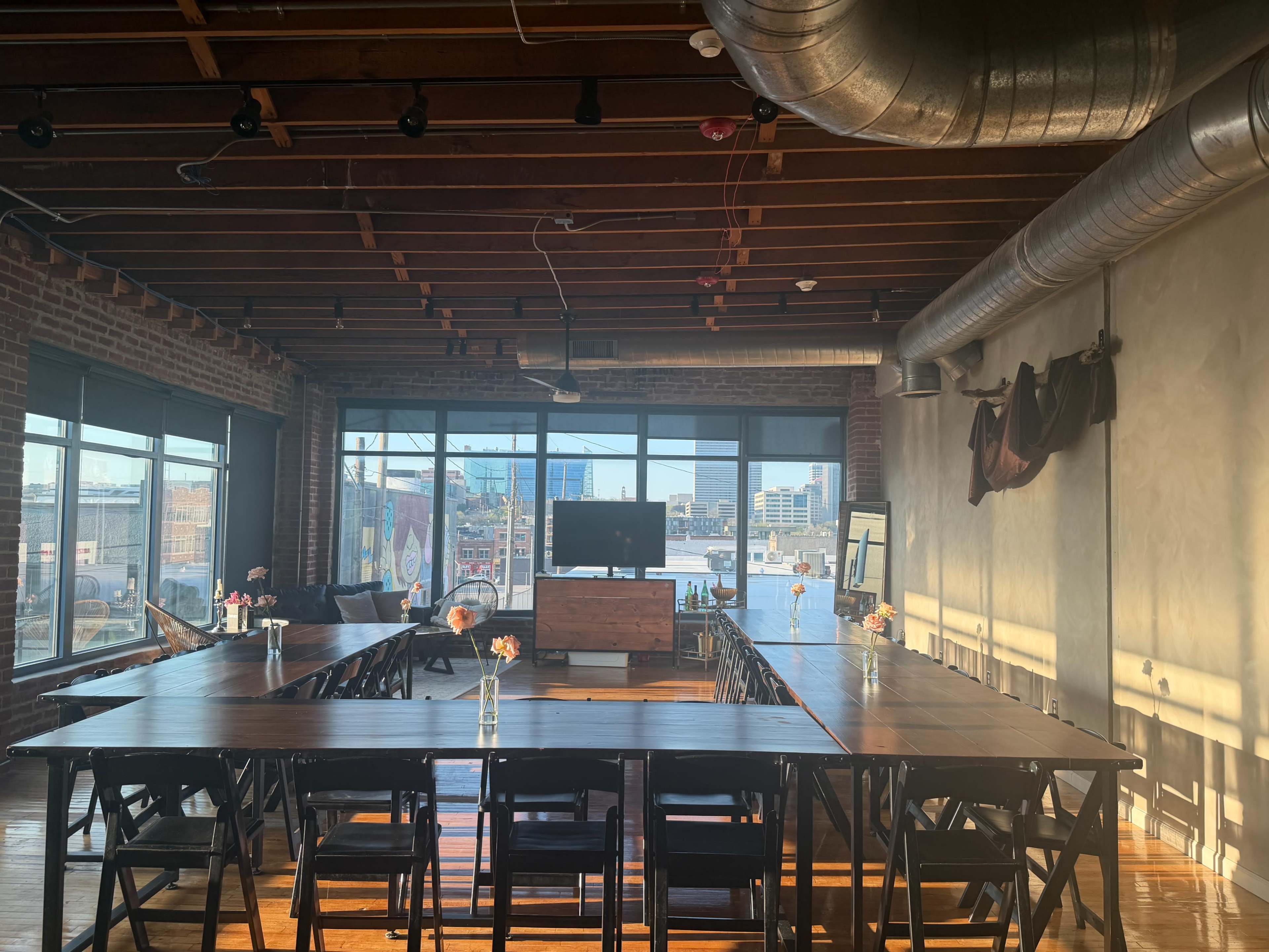 The image shows a spacious meeting room with large windows, a long table set up with chairs on either side, and a television screen mounted on the wall.