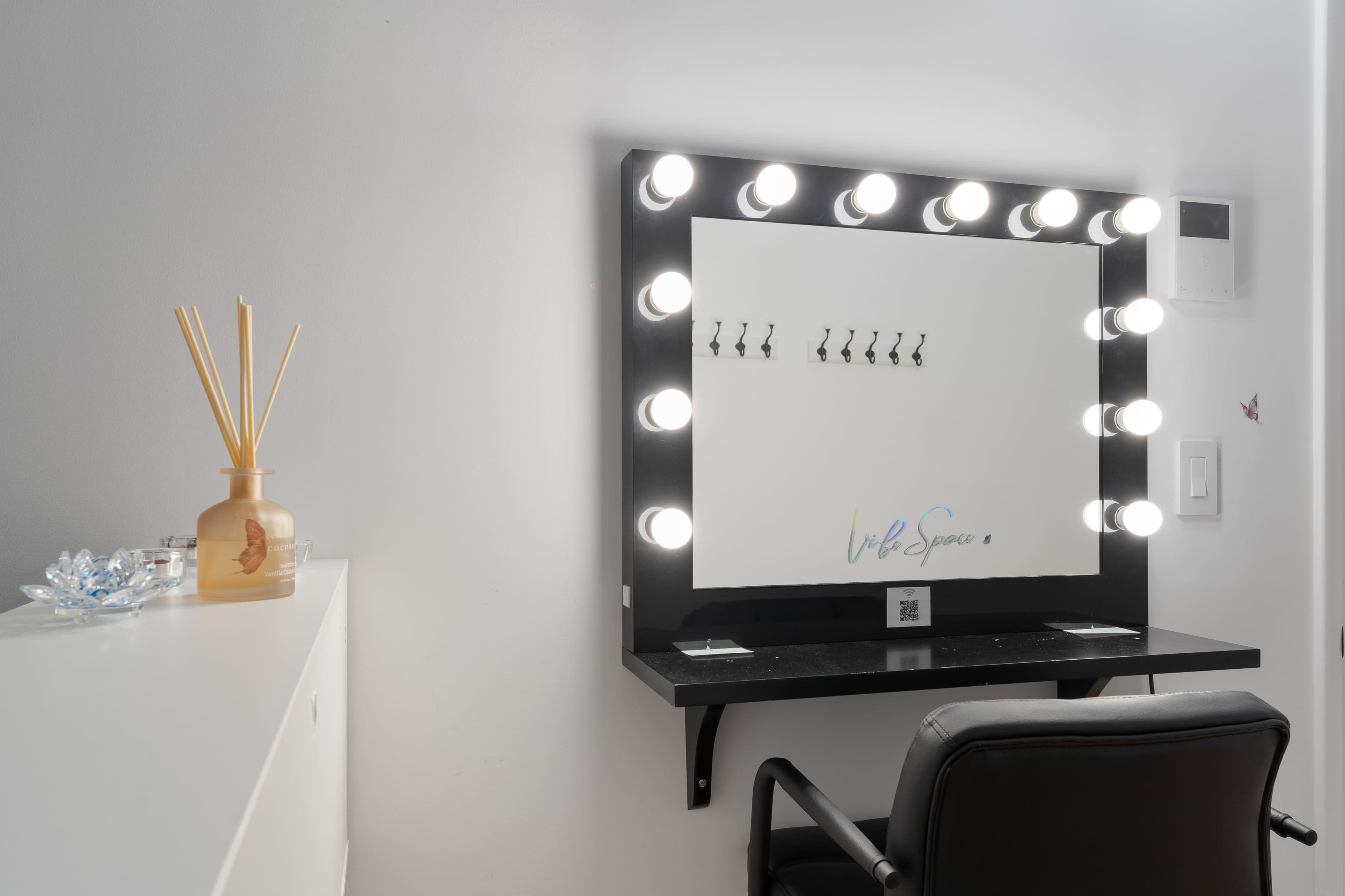 A well-lit vanity mirror with multiple bulbs, a black chair, and a small shelf holding a decorative vase and an ornamental piece.