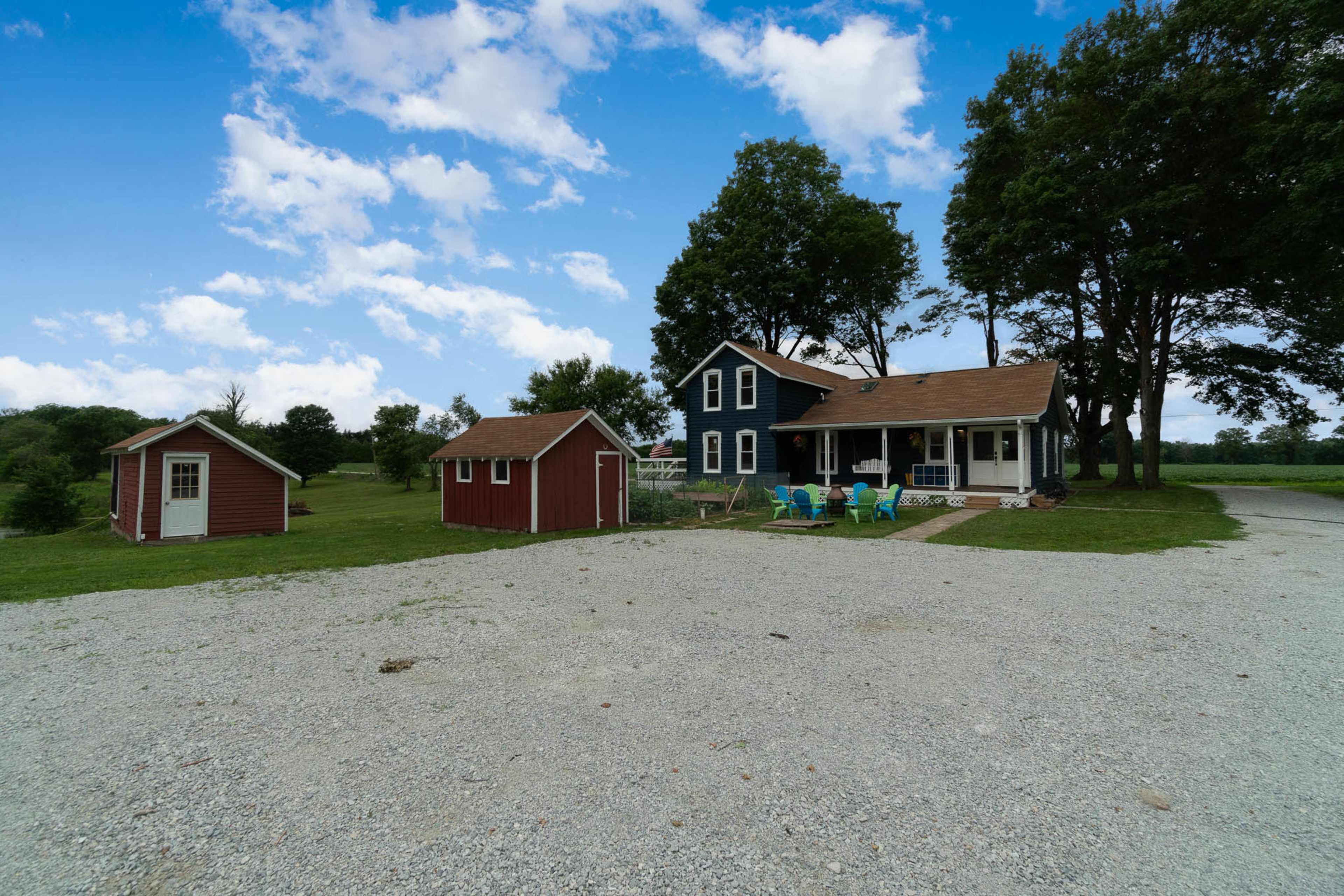 A blue and white farmhouse with an attached porch is situated next to a red shed in a gravel area surrounded by trees under a partly cloudy sky.