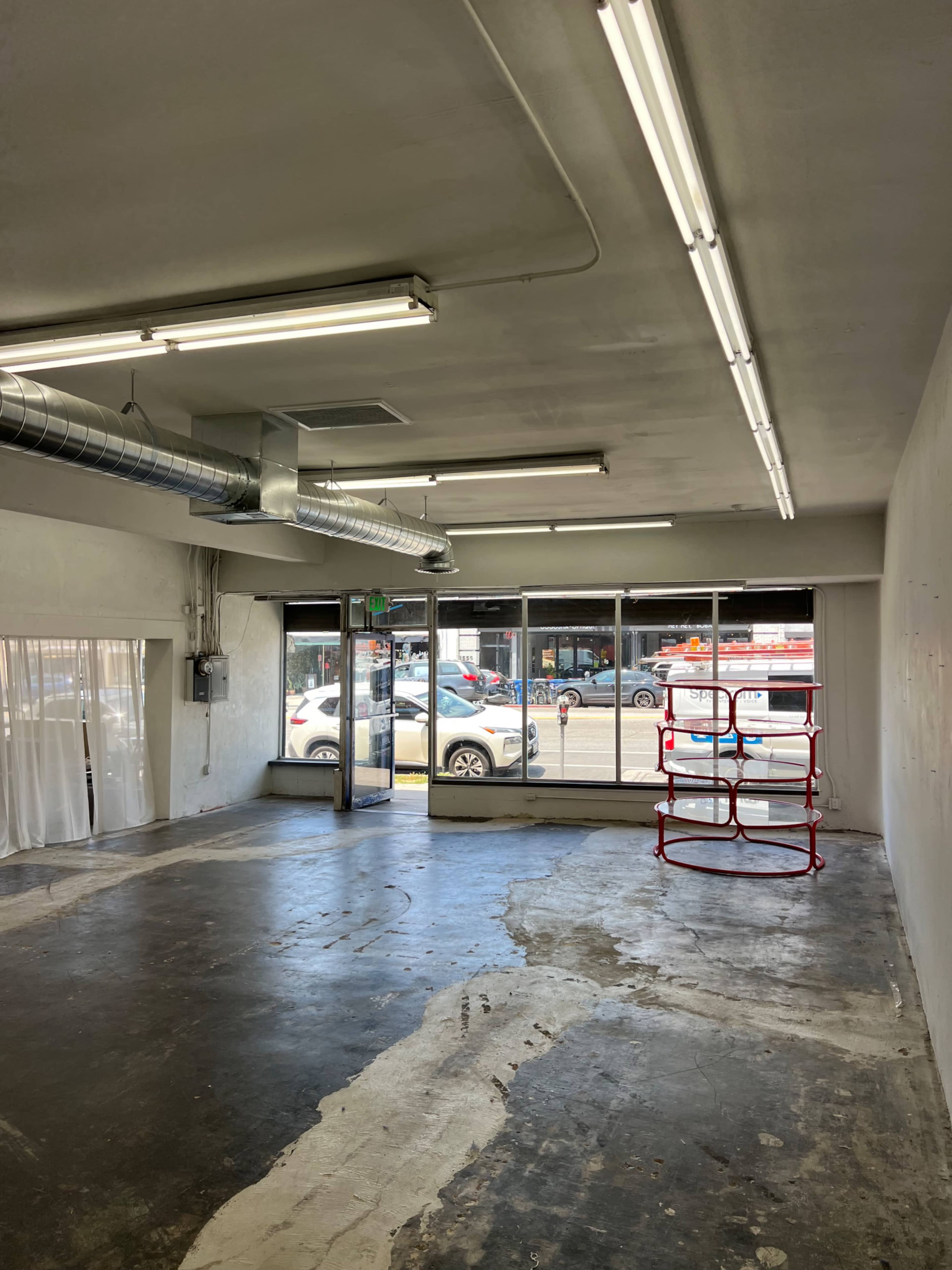 An empty commercial space with a concrete floor, fluorescent lighting, and large windows facing the street.