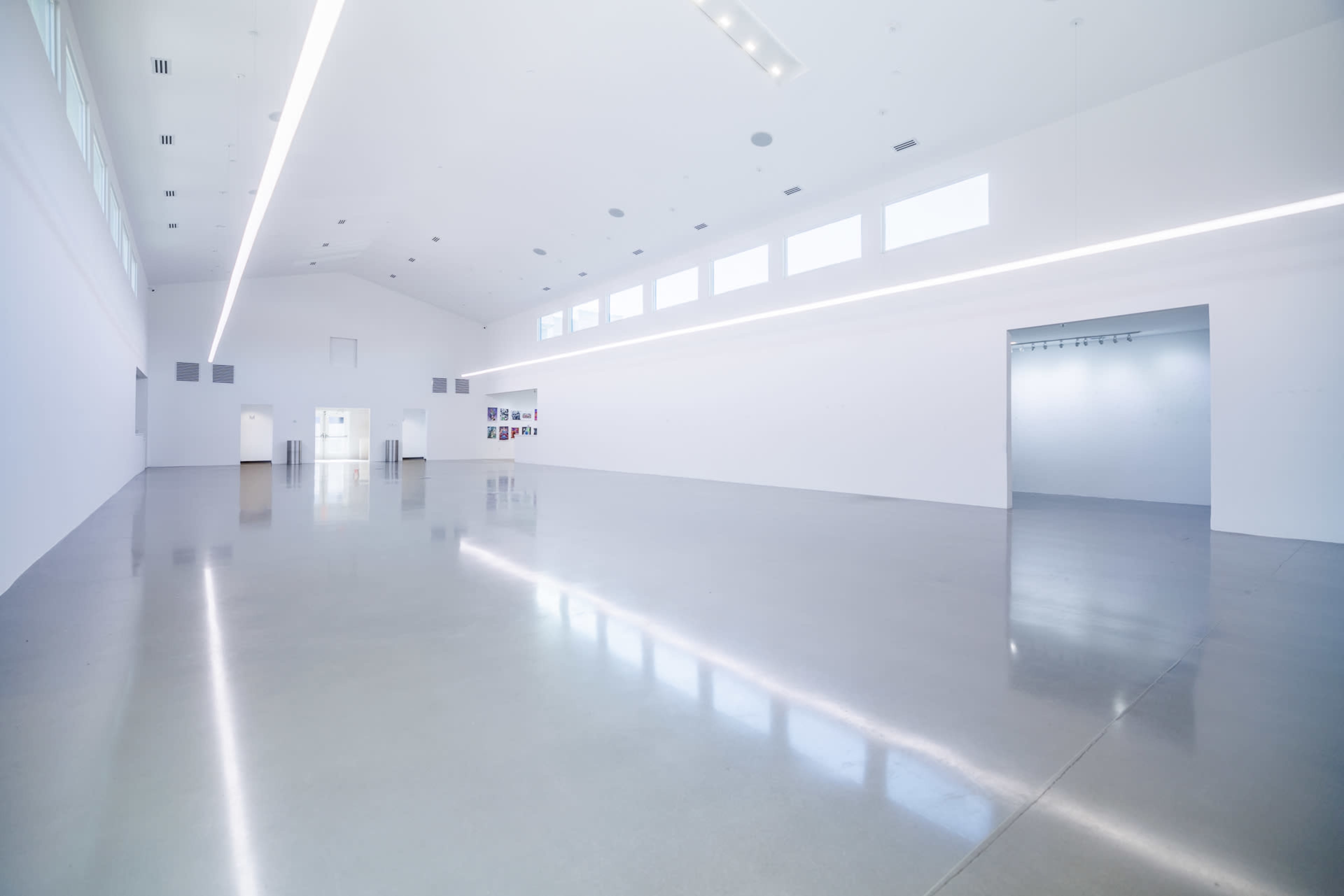 The image shows a spacious, minimalist art gallery with a polished concrete floor and large windows along the upper wall.