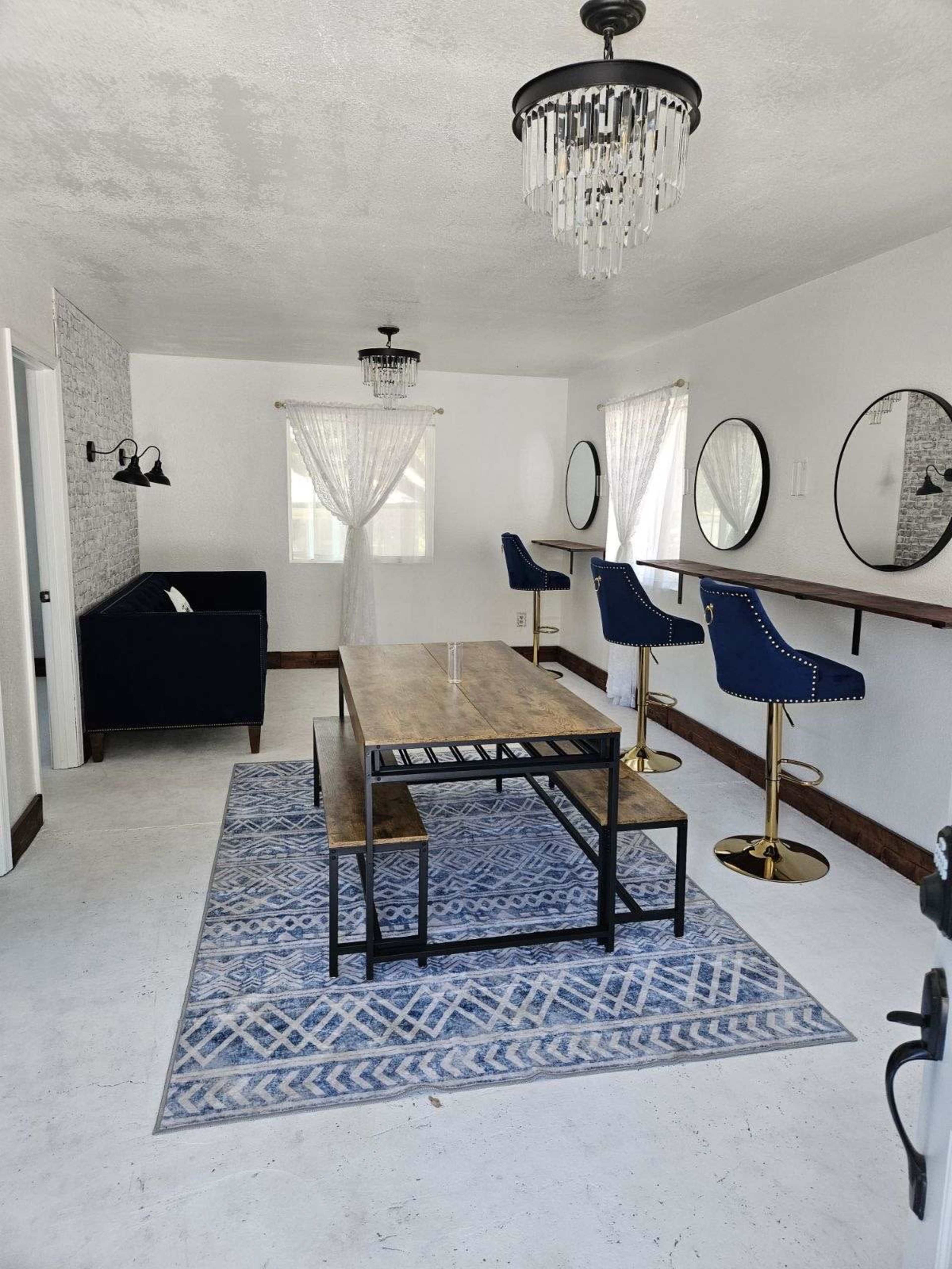 A modern room features a blue couch, two round mirrors, a wooden dining table with benches, and bar stools along a wall, all set on a patterned rug.