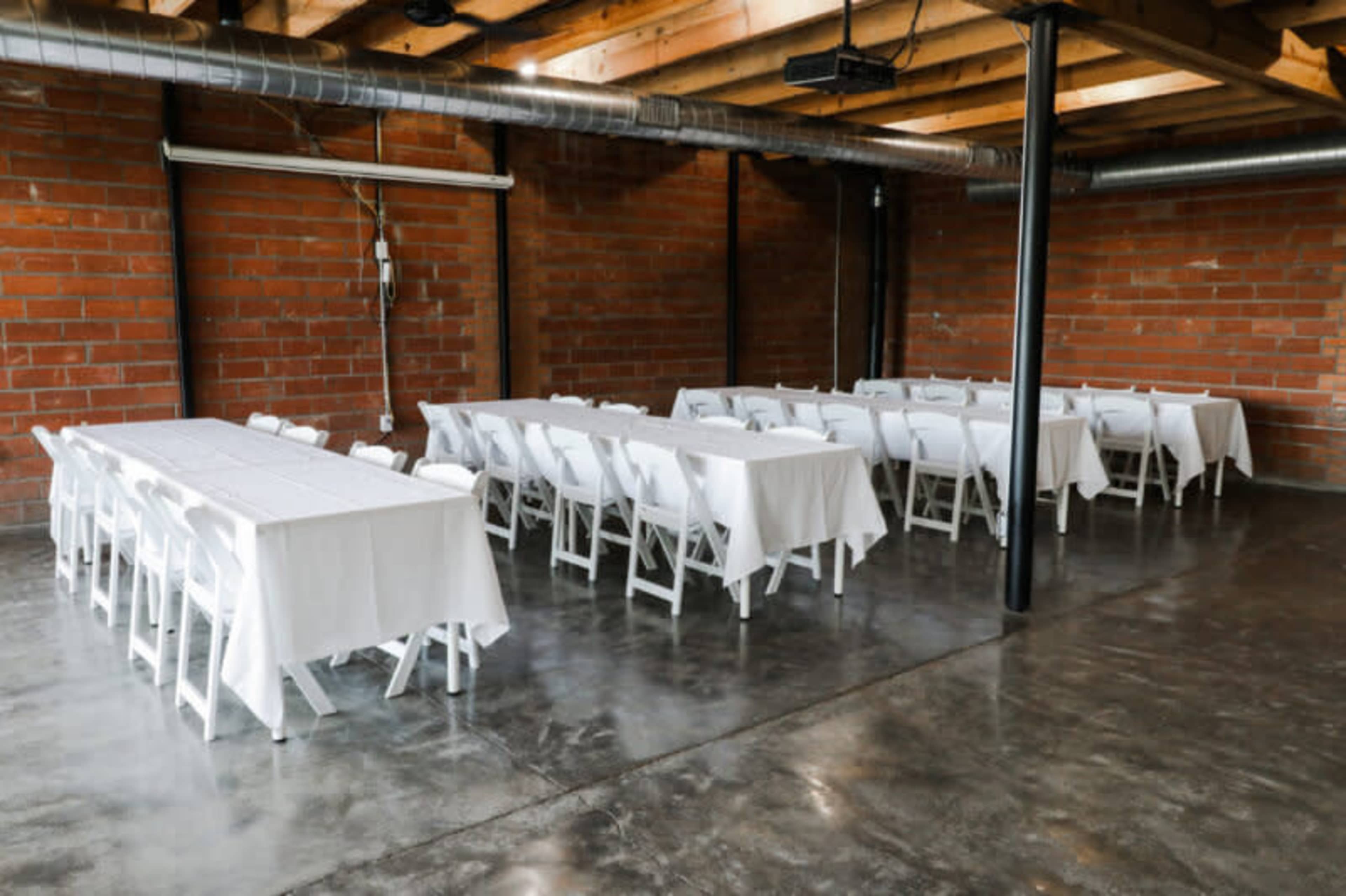 The image shows a spacious industrial room with two long rows of tables covered in white tablecloths and surrounded by white chairs.