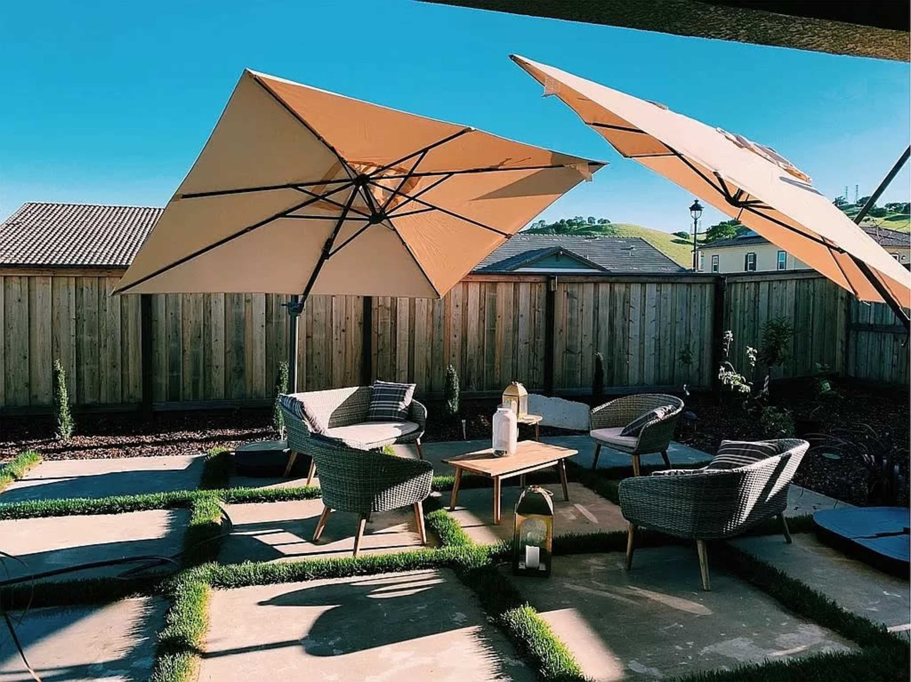 A patio area features two large umbrellas over a seating arrangement of wicker chairs and a wooden table, surrounded by grass and a wooden fence.