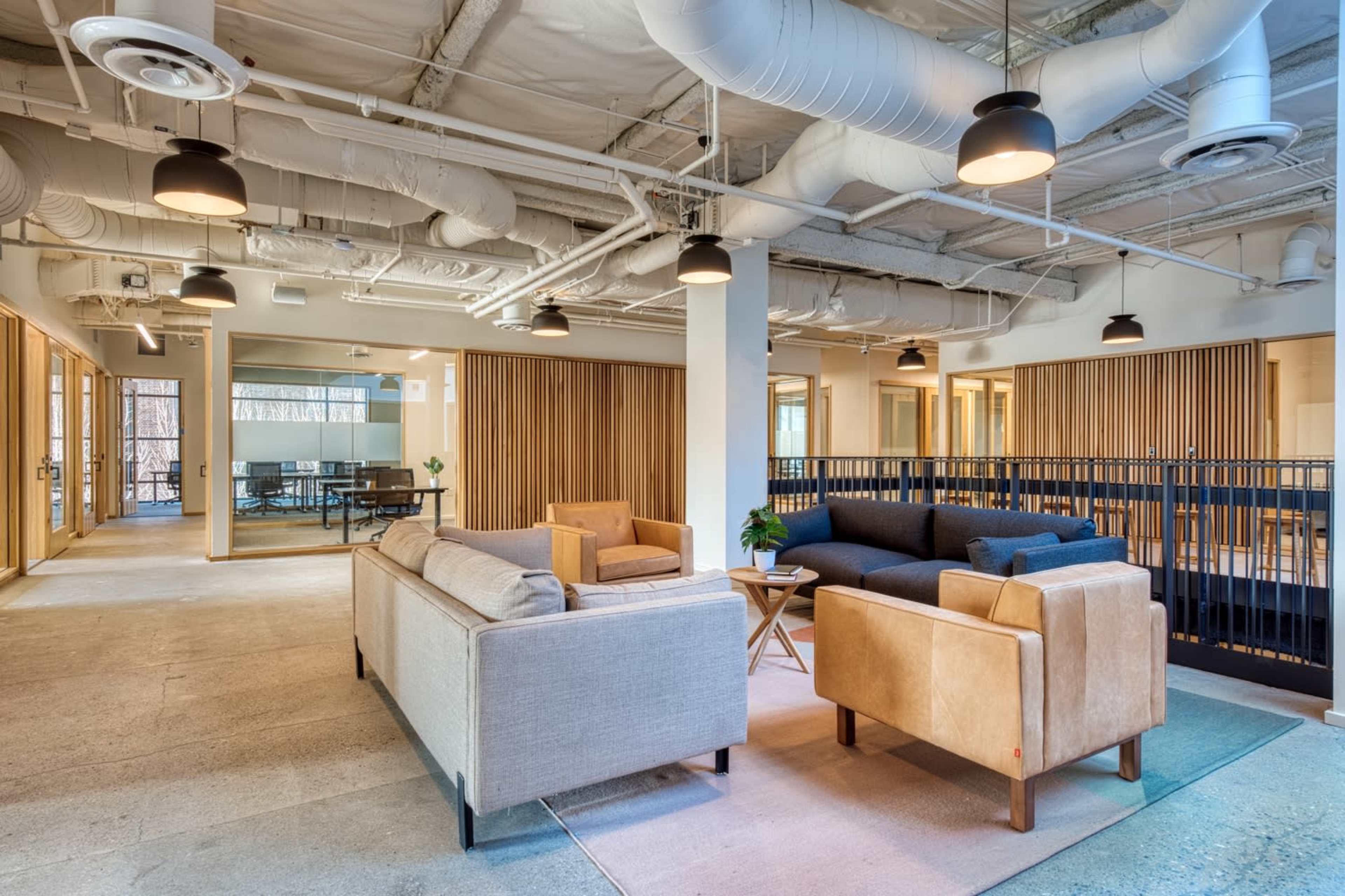 A modern open-plan office space featuring seating areas with gray and tan sofas, wooden partitions, and large windows.
