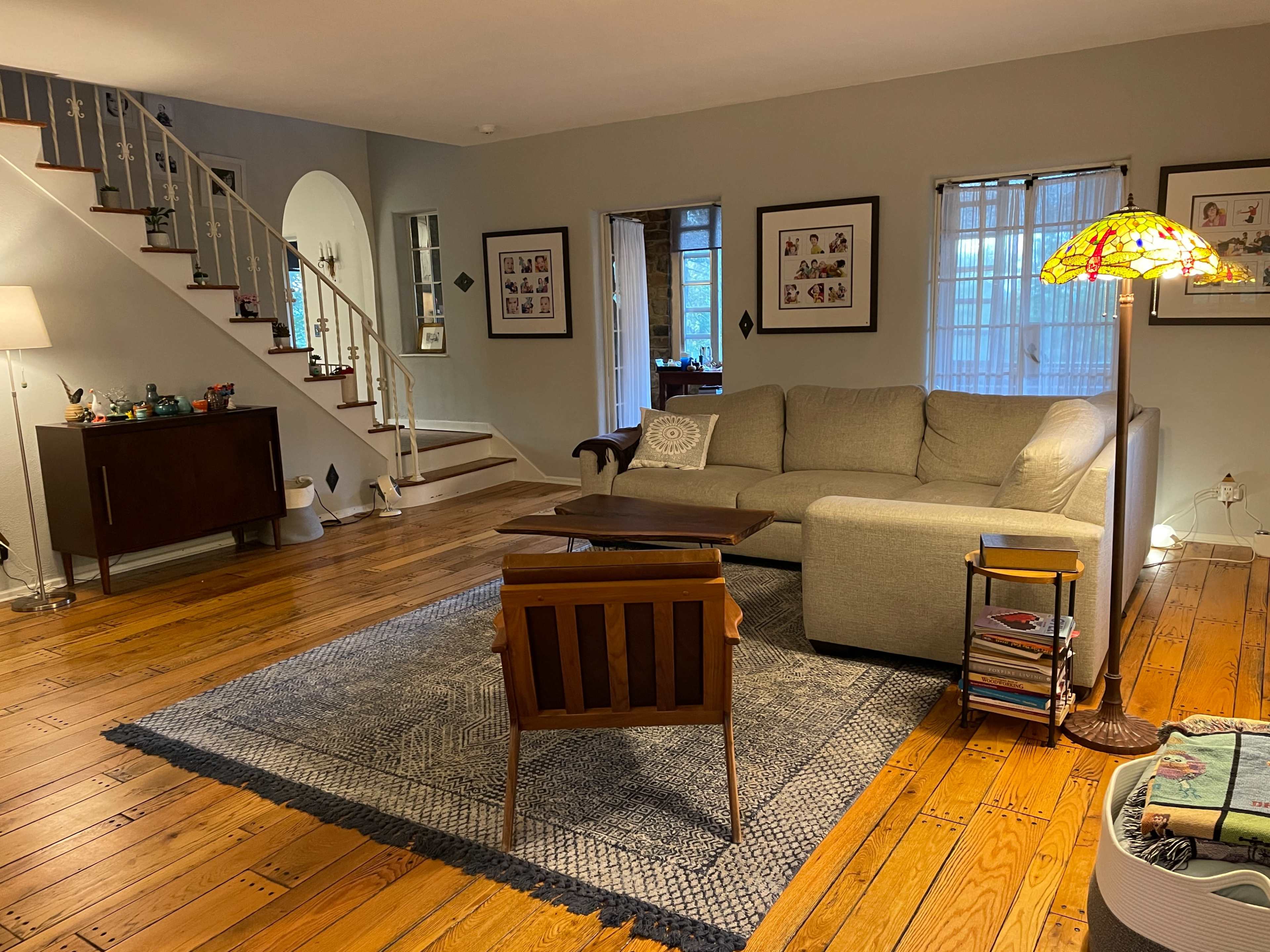 The image shows a cozy living room with a sectional sofa, wooden coffee table, and a decorative lamp, featuring hardwood floors and a staircase in the background.