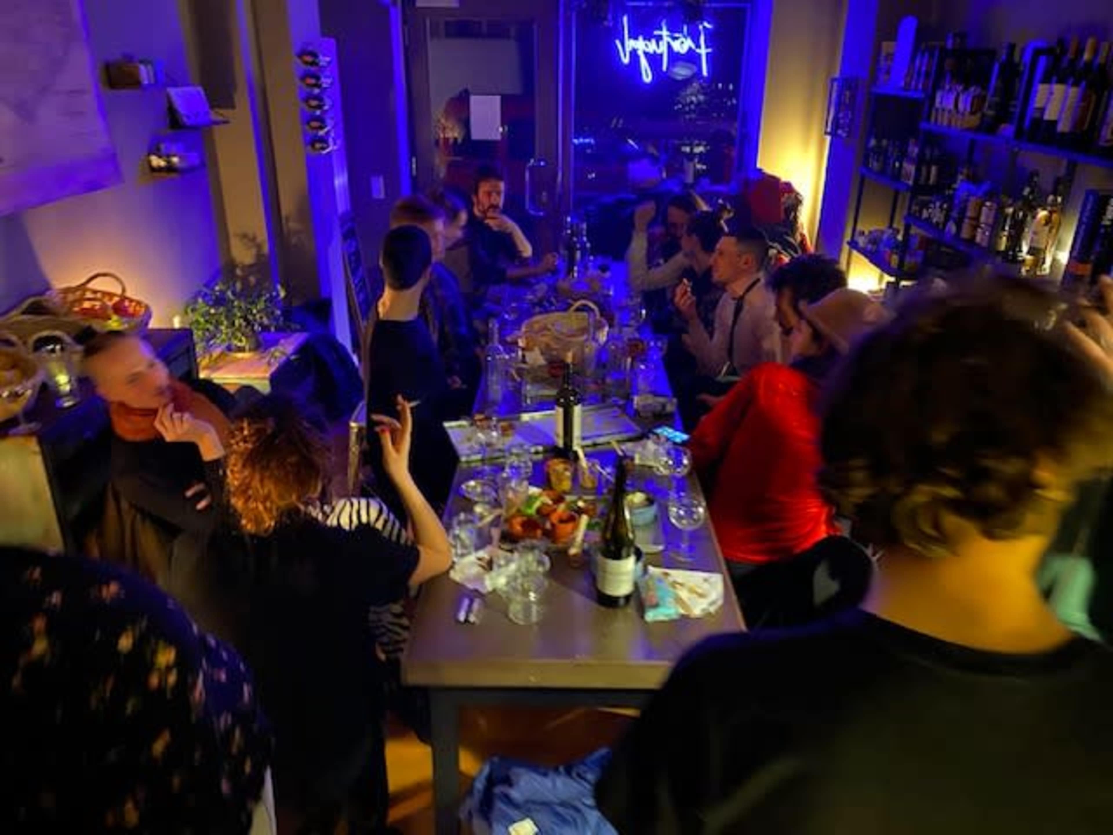 A group of people gathers around a long table in a dimly lit room filled with bottles and city views at night.
