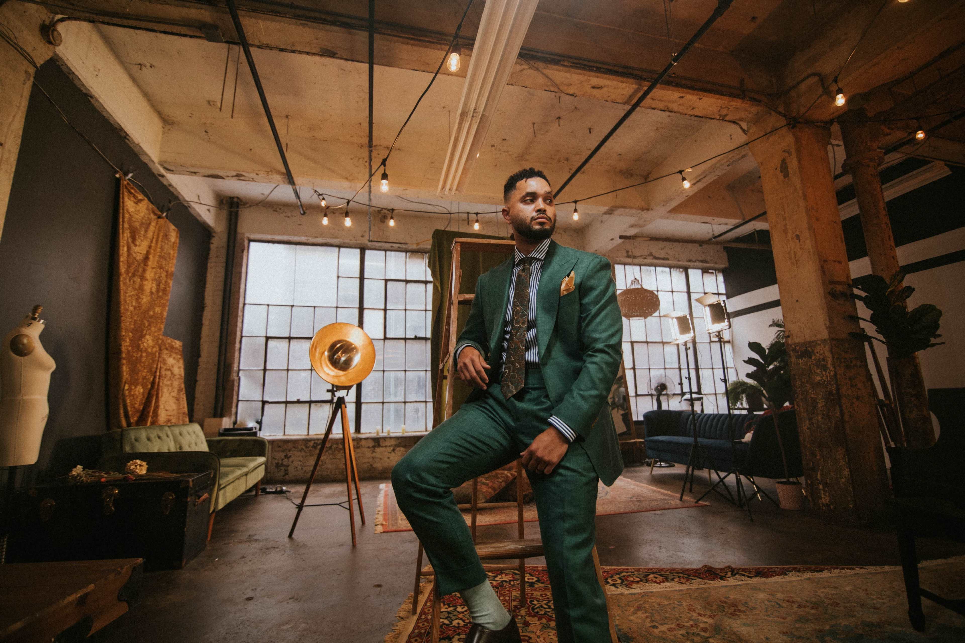 A man in a green suit poses confidently in a stylish industrial studio with large windows and decorative lighting.