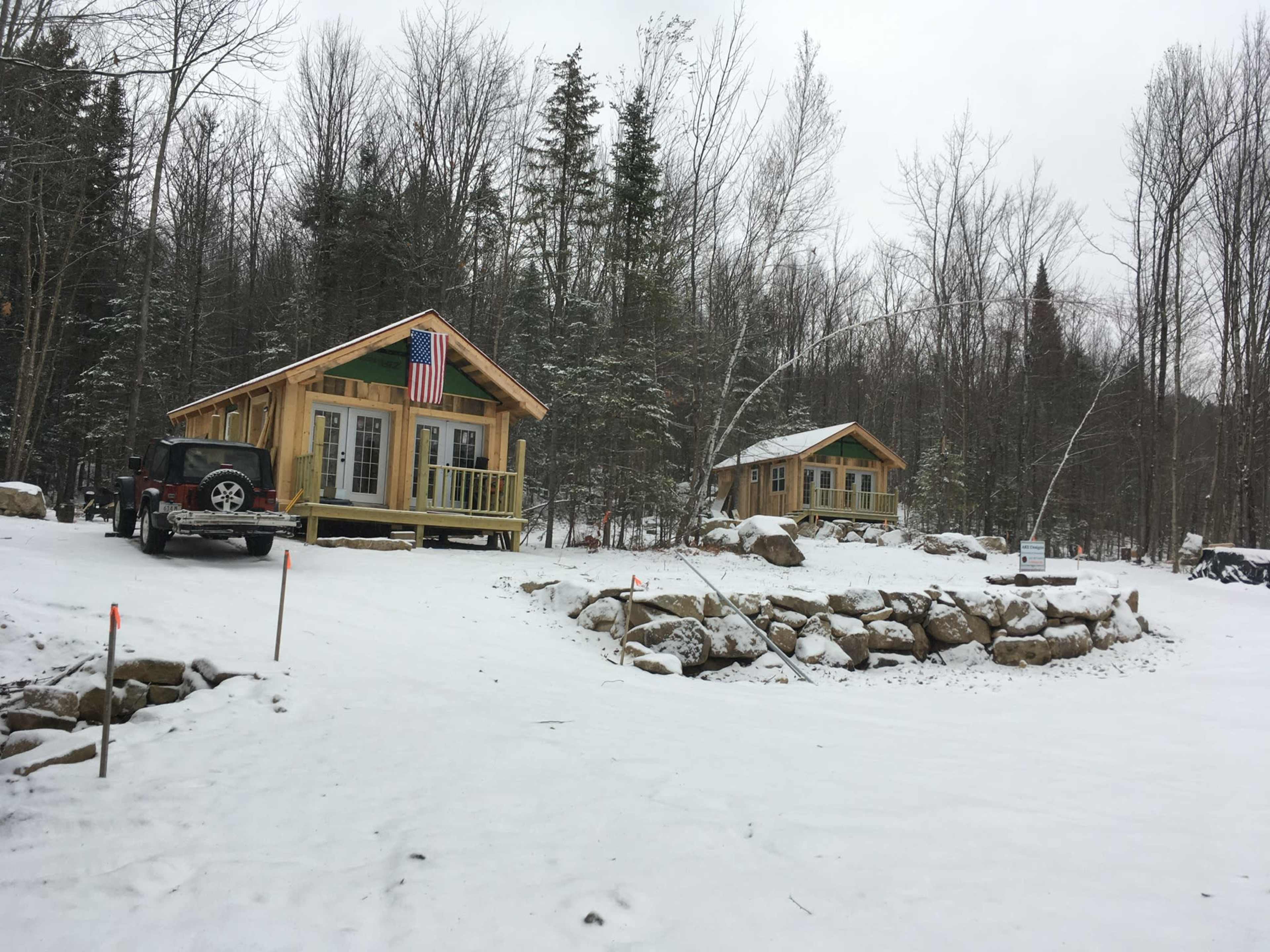 Two wooden cabins are situated on a snowy landscape surrounded by trees, with an American flag displayed on one of the cabins.