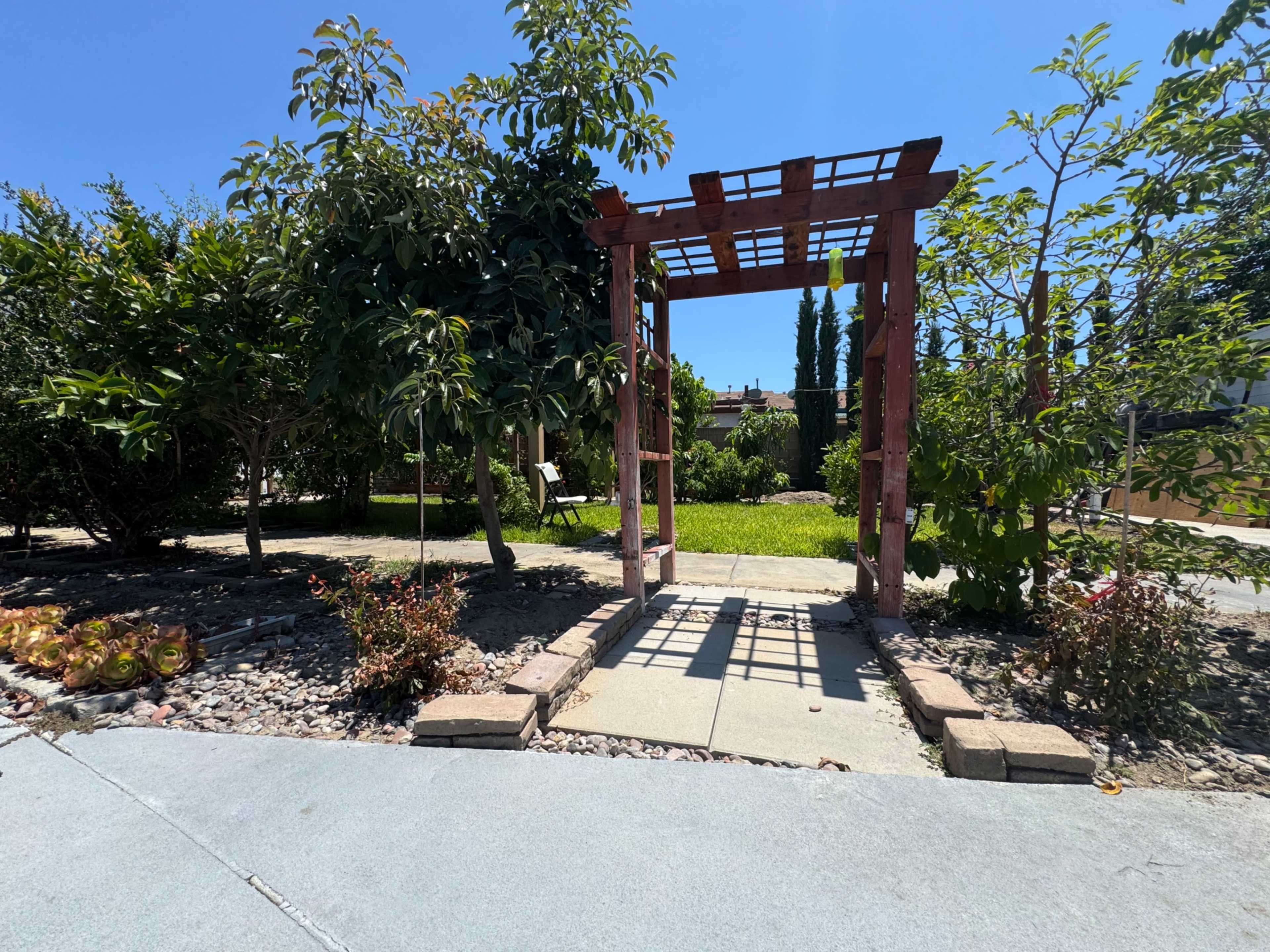 A wooden arbor surrounded by greenery leads into a grassy area, with a pathway made of stone pavers visible in the foreground.