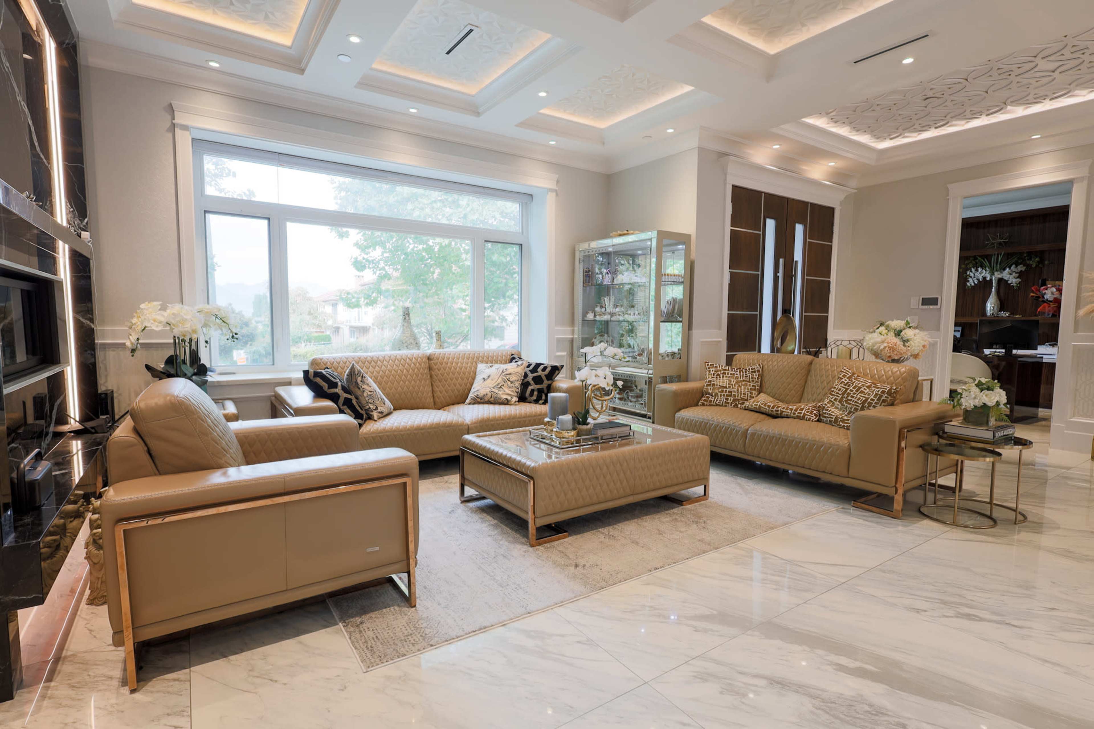 The image shows a modern living room with light-colored leather sofas, a marble floor, and a large window that lets in natural light.