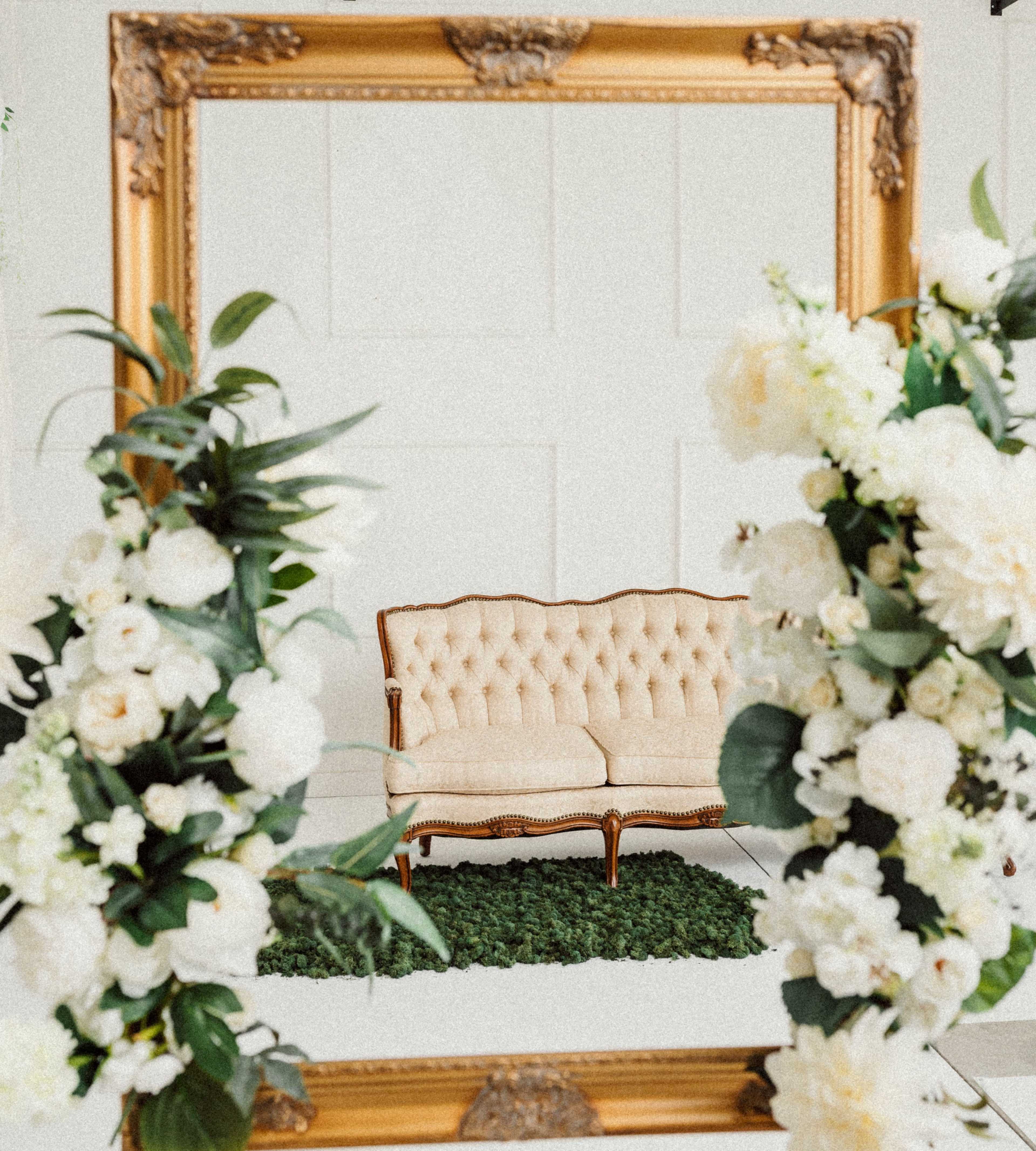 A vintage gold-framed mirror reflects a cream-colored loveseat surrounded by white floral arrangements.