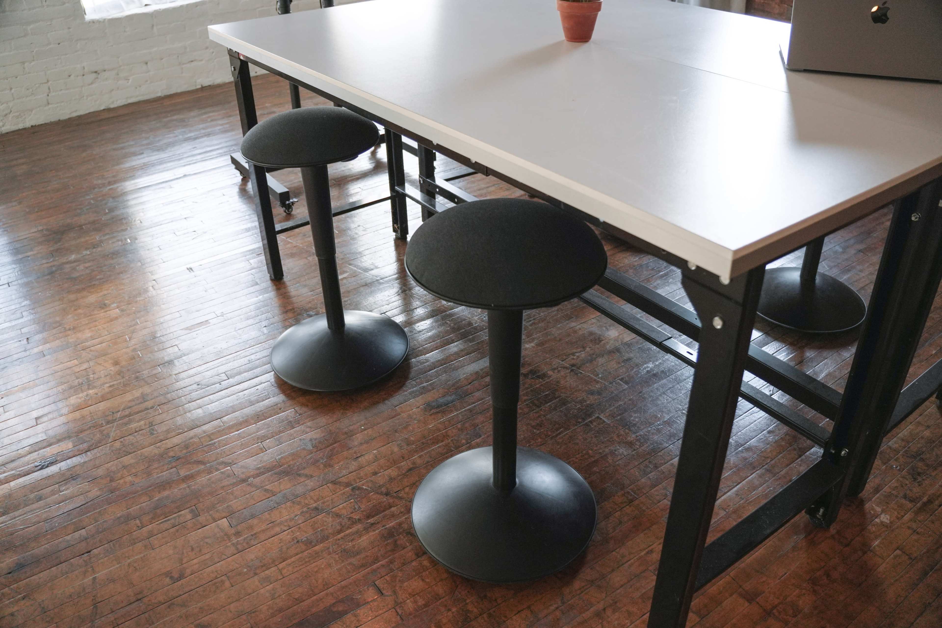 A modern workspace features a white table with two black stools on a wooden floor.