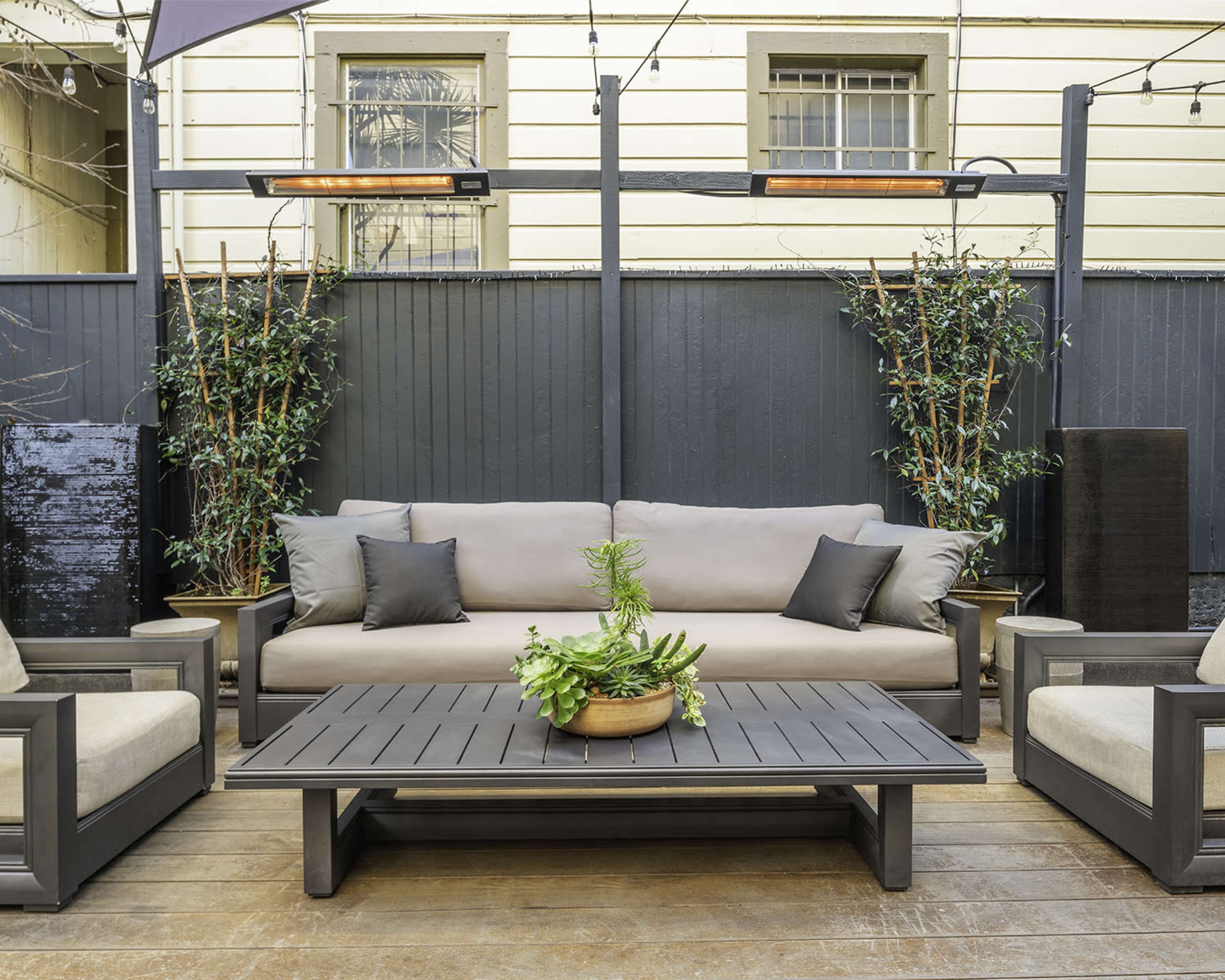 The image shows a patio area featuring a gray couch with pillows, a wooden coffee table with a plant centerpiece, and two armchairs, all surrounded by potted greenery and a wooden fence.
