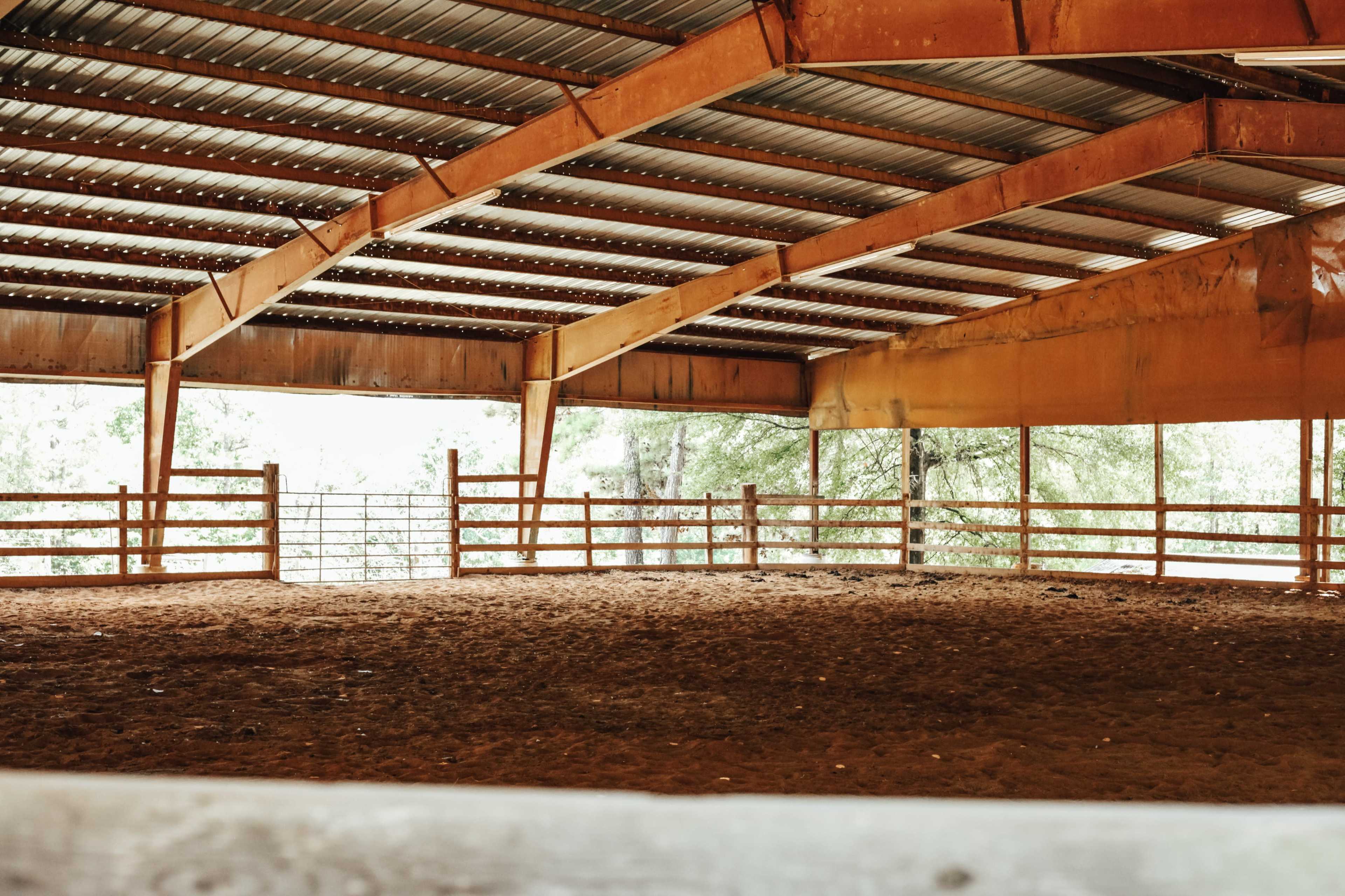 The image shows a large indoor riding arena with a dirt floor and wooden fencing under a metal roof.