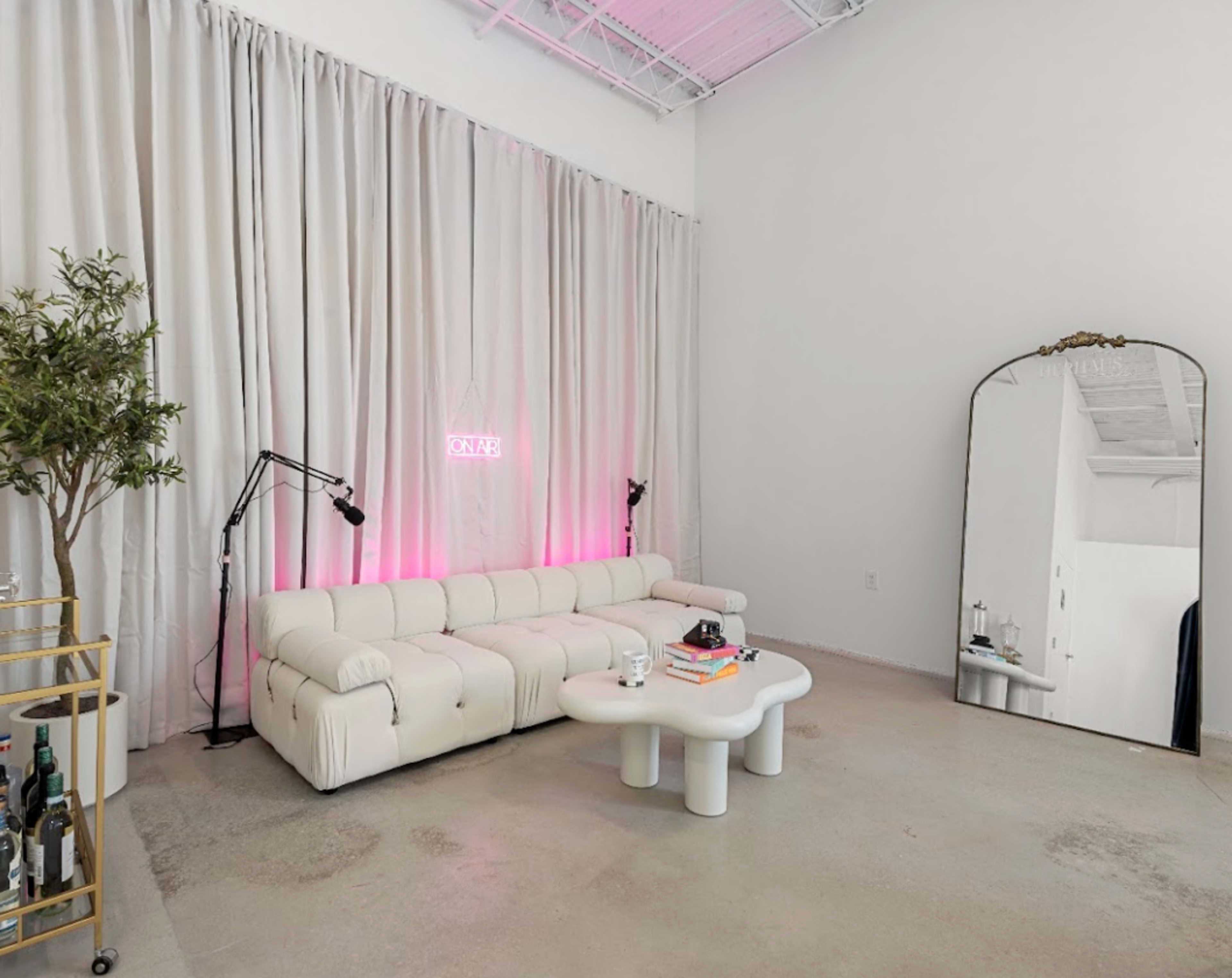 The scene features a minimalist living room with a beige sofa, a white coffee table, a tall mirror, and pink neon lights against a backdrop of cream curtains.