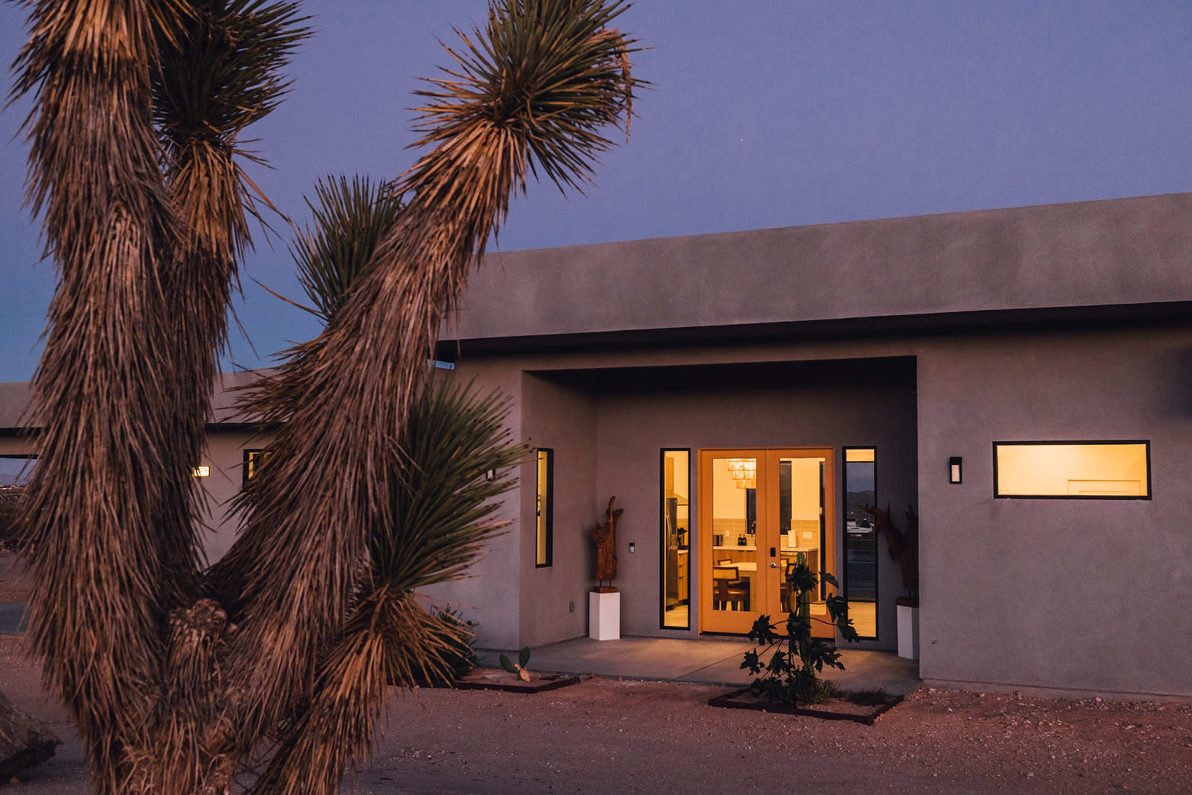 A modern building with large windows is illuminated at dusk, surrounded by desert plants.
