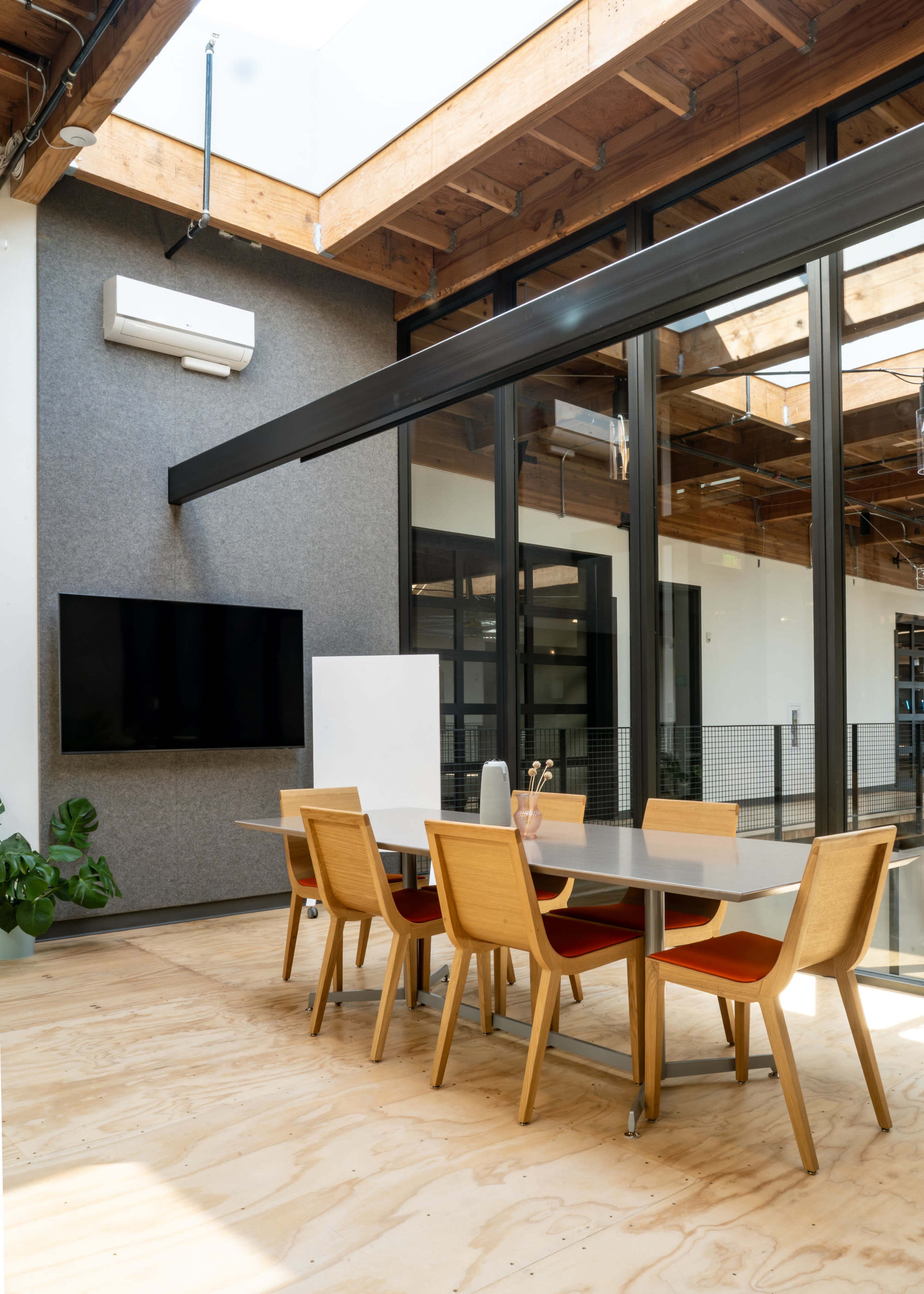 A modern conference room features a large table surrounded by wooden chairs, with a television mounted on the wall and large windows allowing natural light to enter.