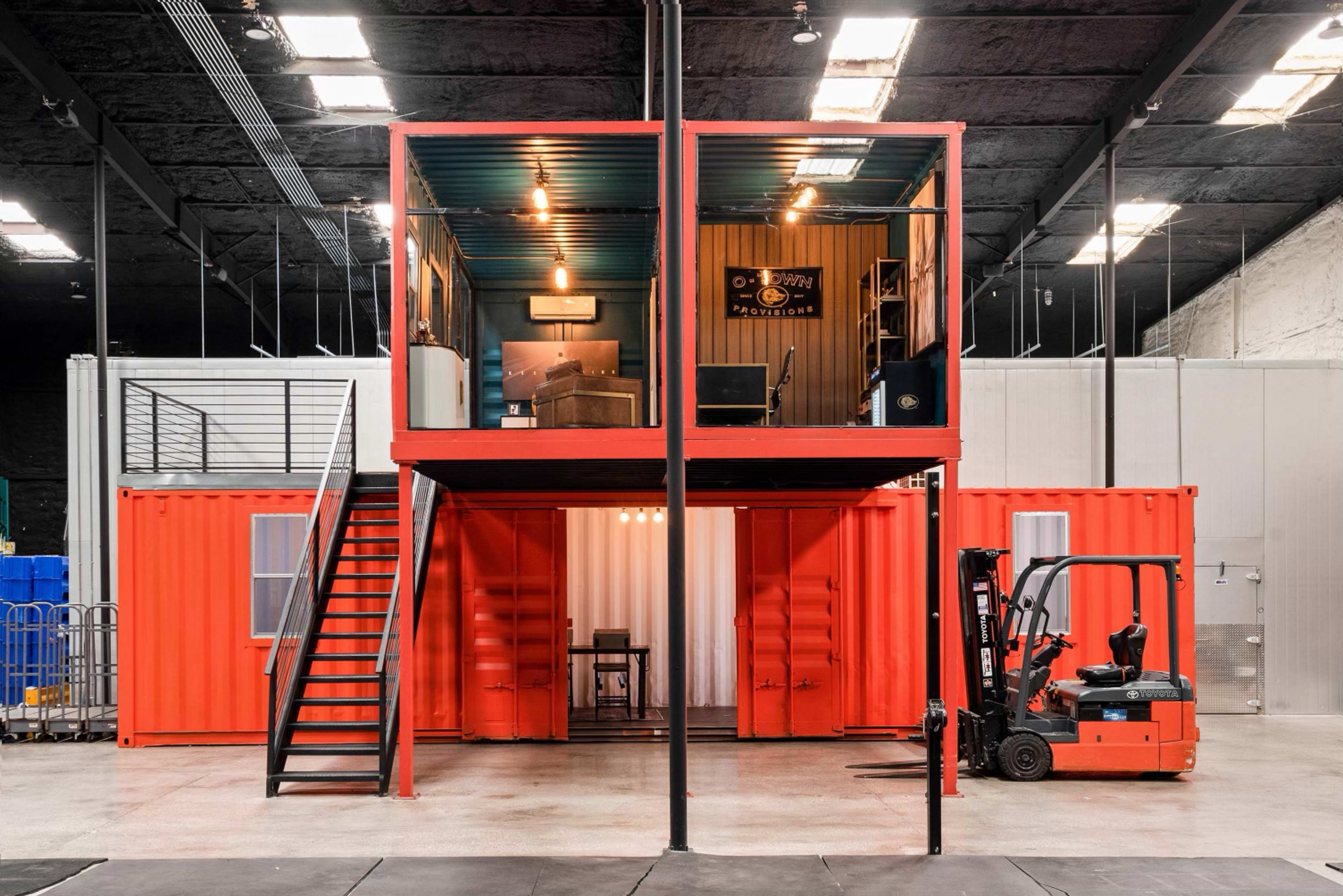 The image shows a two-level structure made from shipping containers, featuring a staircase and industrial lighting in an interior space.