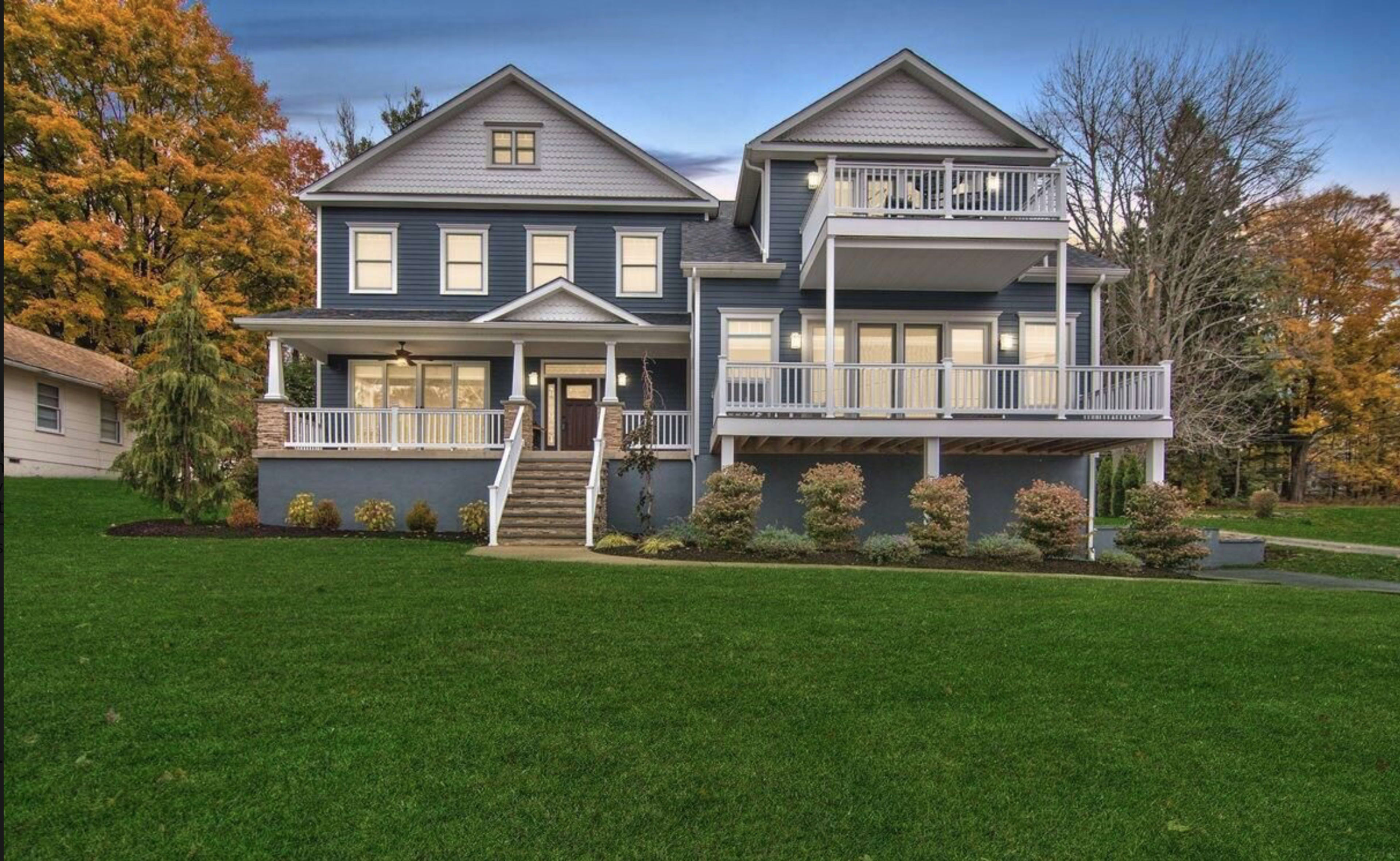 A large, two-story blue house with a white porch and multiple balconies, surrounded by a neatly manicured lawn and trees in autumn foliage.
