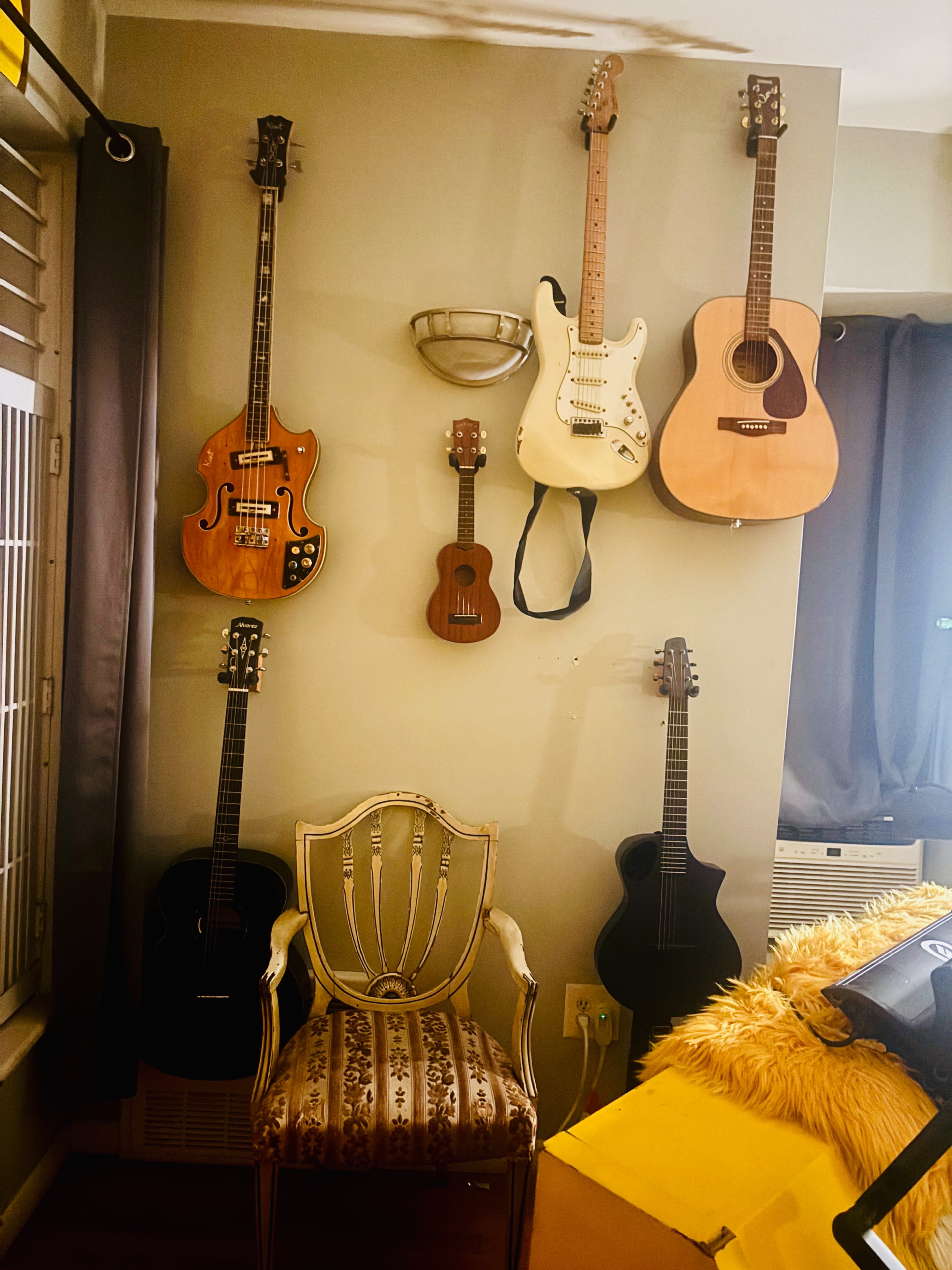 The image shows a wall adorned with various guitars and a ukulele, alongside a decorative chair positioned in front of it.