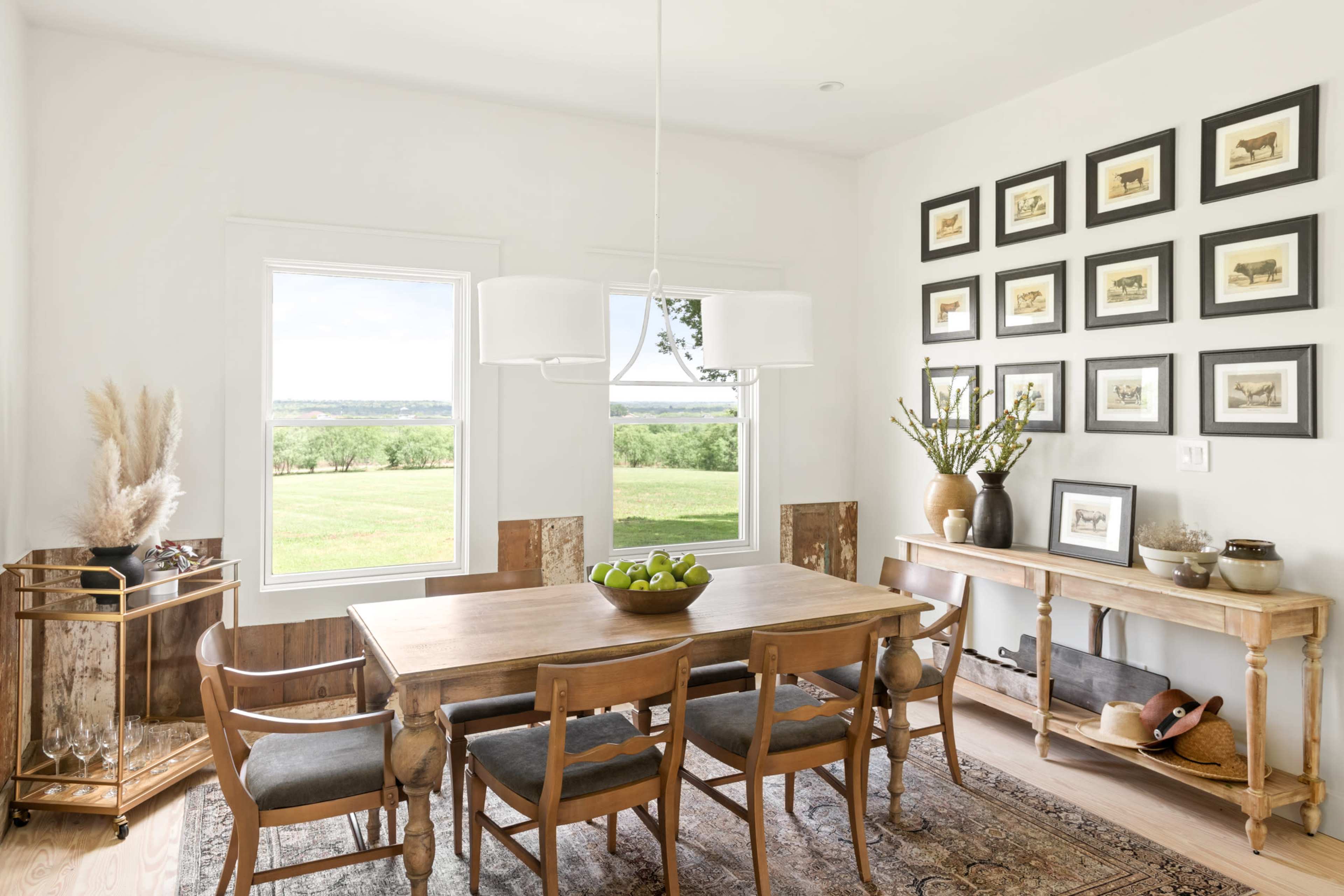 A bright dining room features a wooden table with four chairs, surrounded by large windows and framed artwork on the walls.