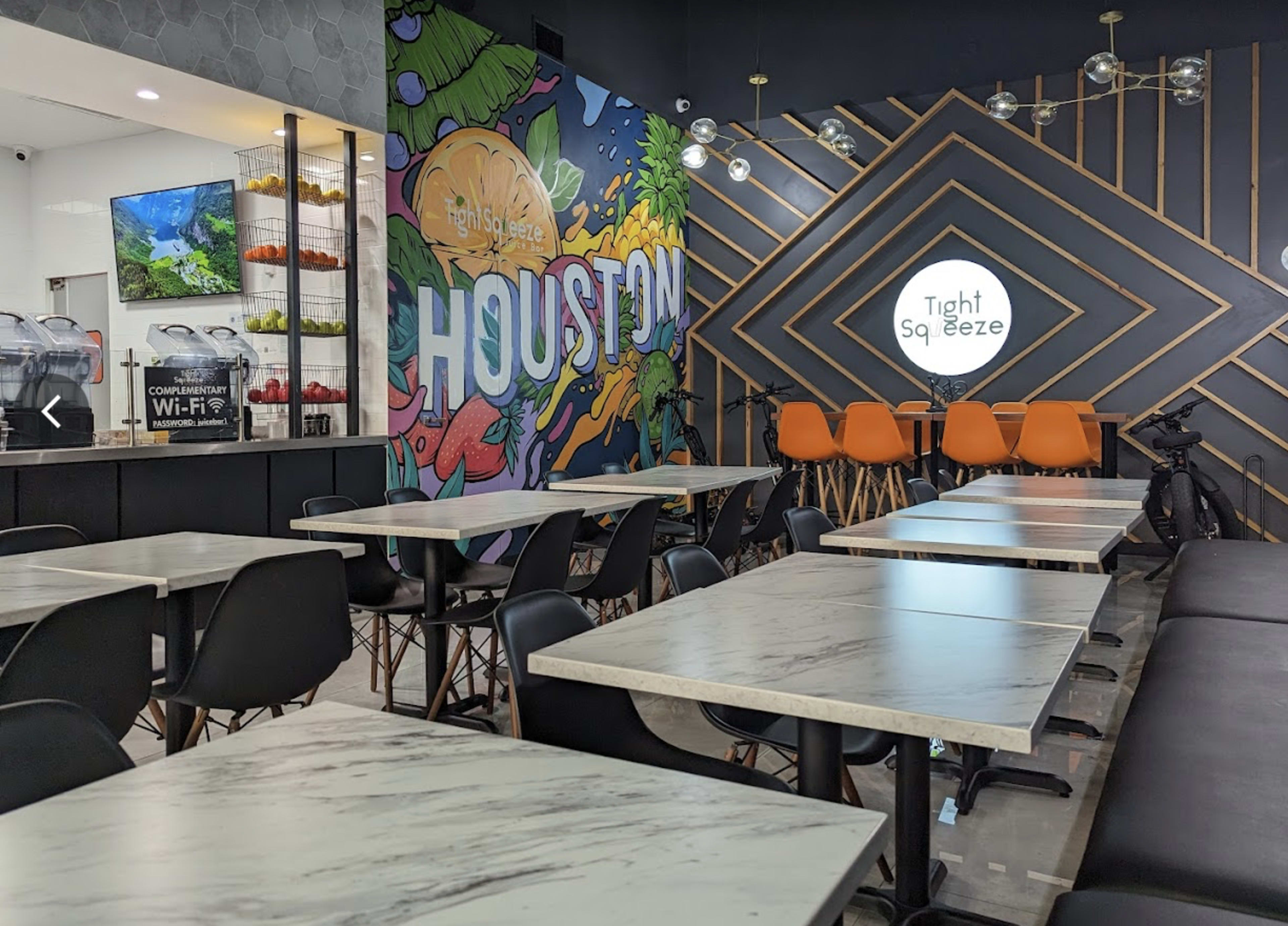 The image shows a modern café interior with marble-topped tables, black chairs, and a colorful mural featuring the word "HOUSTON" on the wall, alongside orange seating and a service counter.