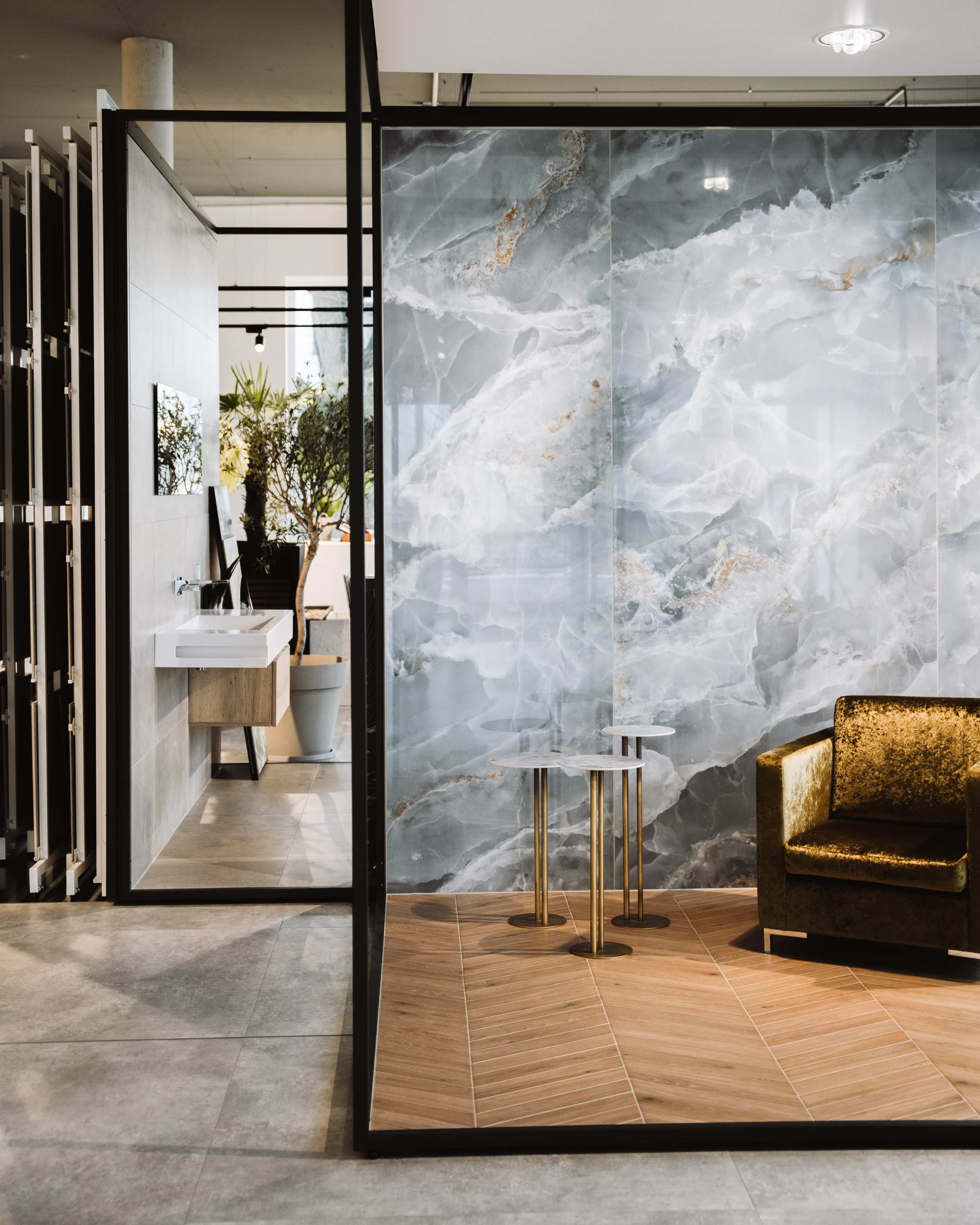 The image shows a modern interior with a gray marble wall, a small wooden floor area, a gold-accented chair, and a view of a sink and plants beyond a glass partition.