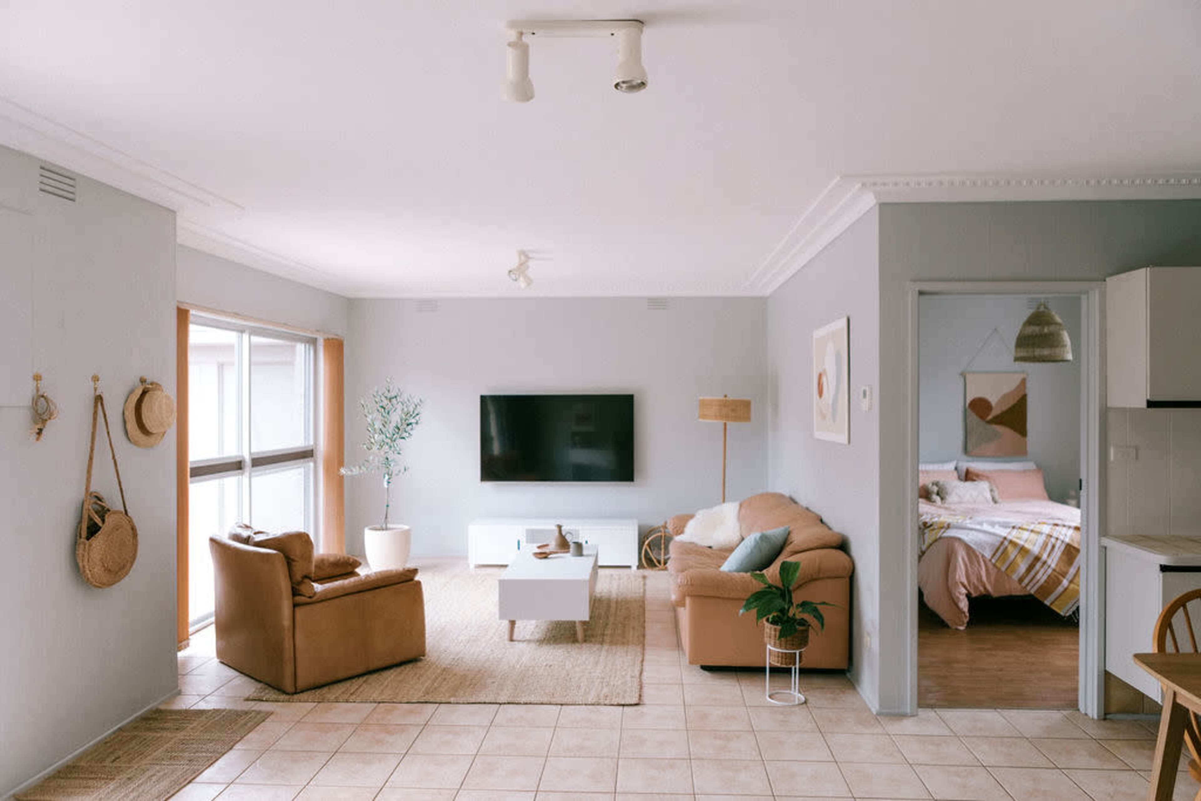 A well-lit, modern living room with a large television, comfortable seating, and a neutral color palette, featuring an adjacent kitchen area and bedroom.