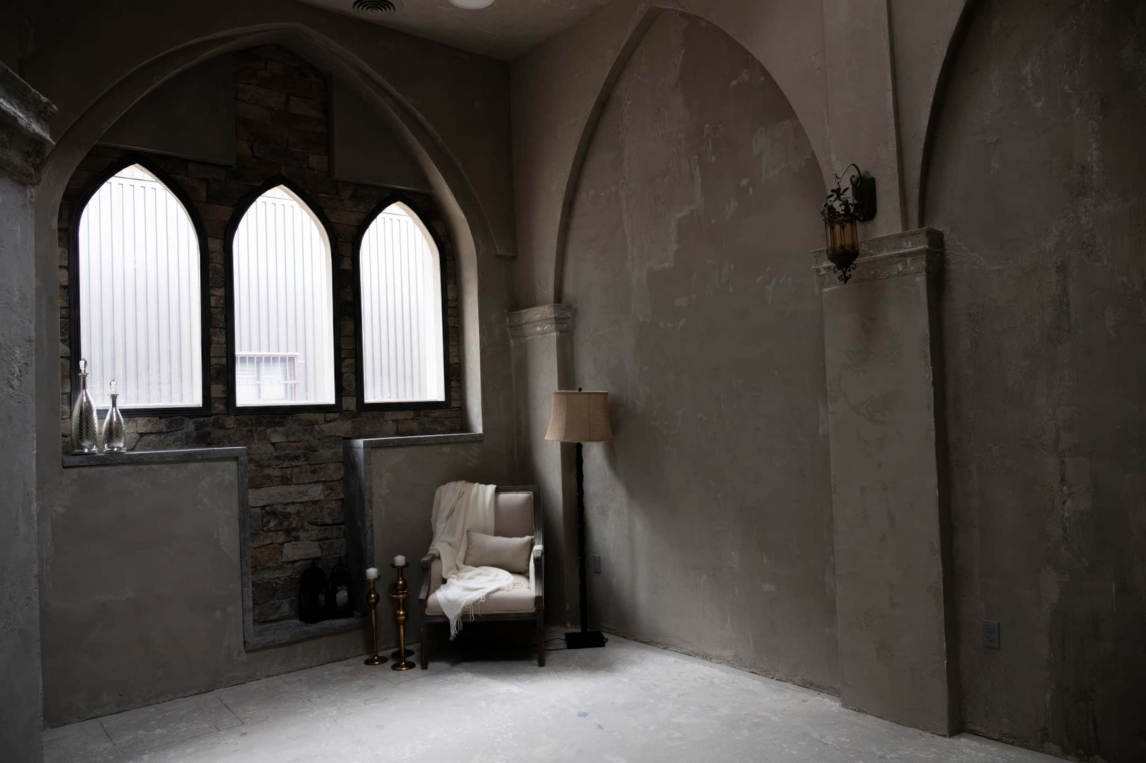 The image shows an empty room with arched windows, bare stone walls, and a single chair beside a lamp.