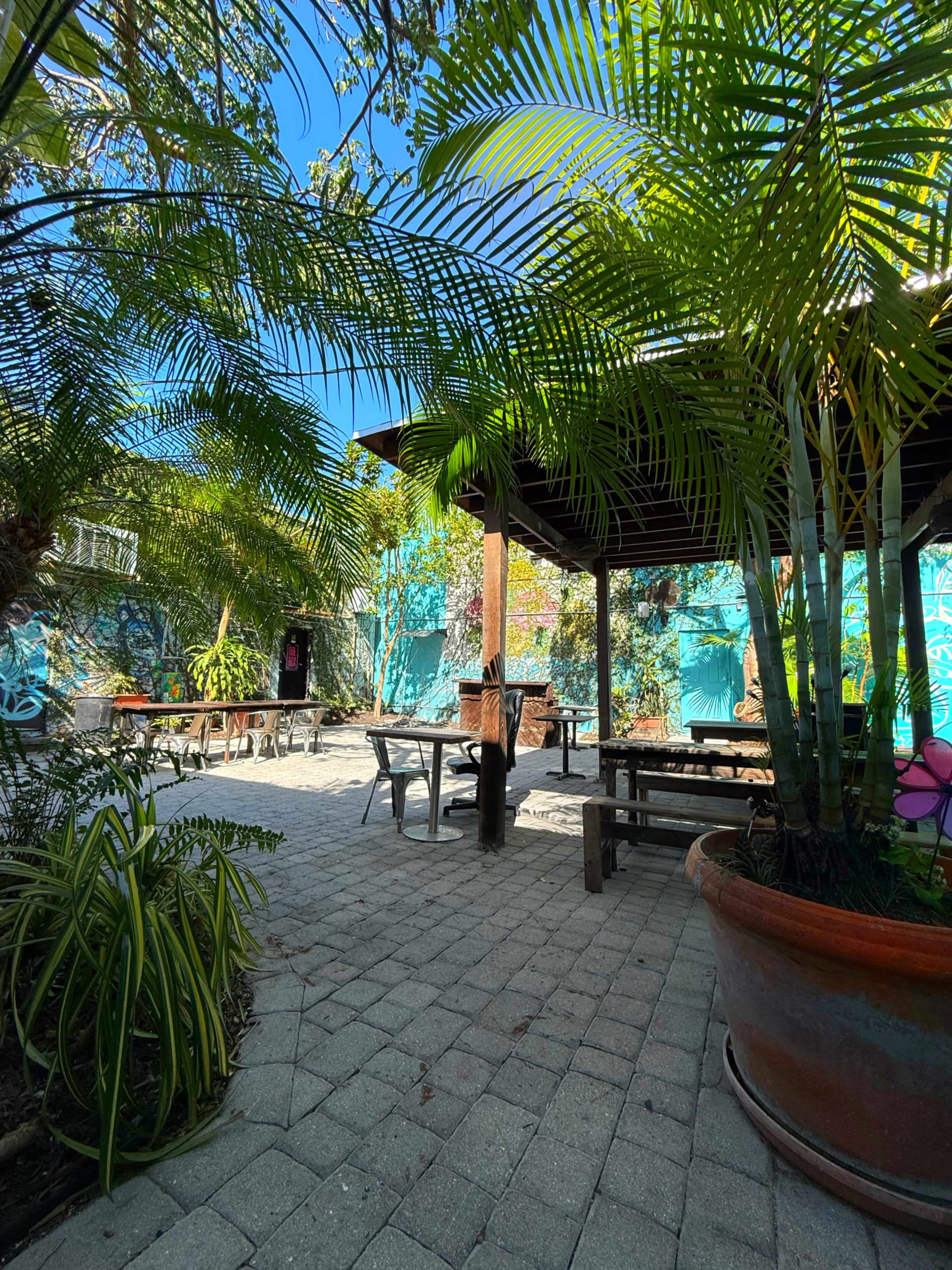 The image shows a courtyard with stone pathways, surrounded by lush greenery and decorated walls featuring colorful murals.