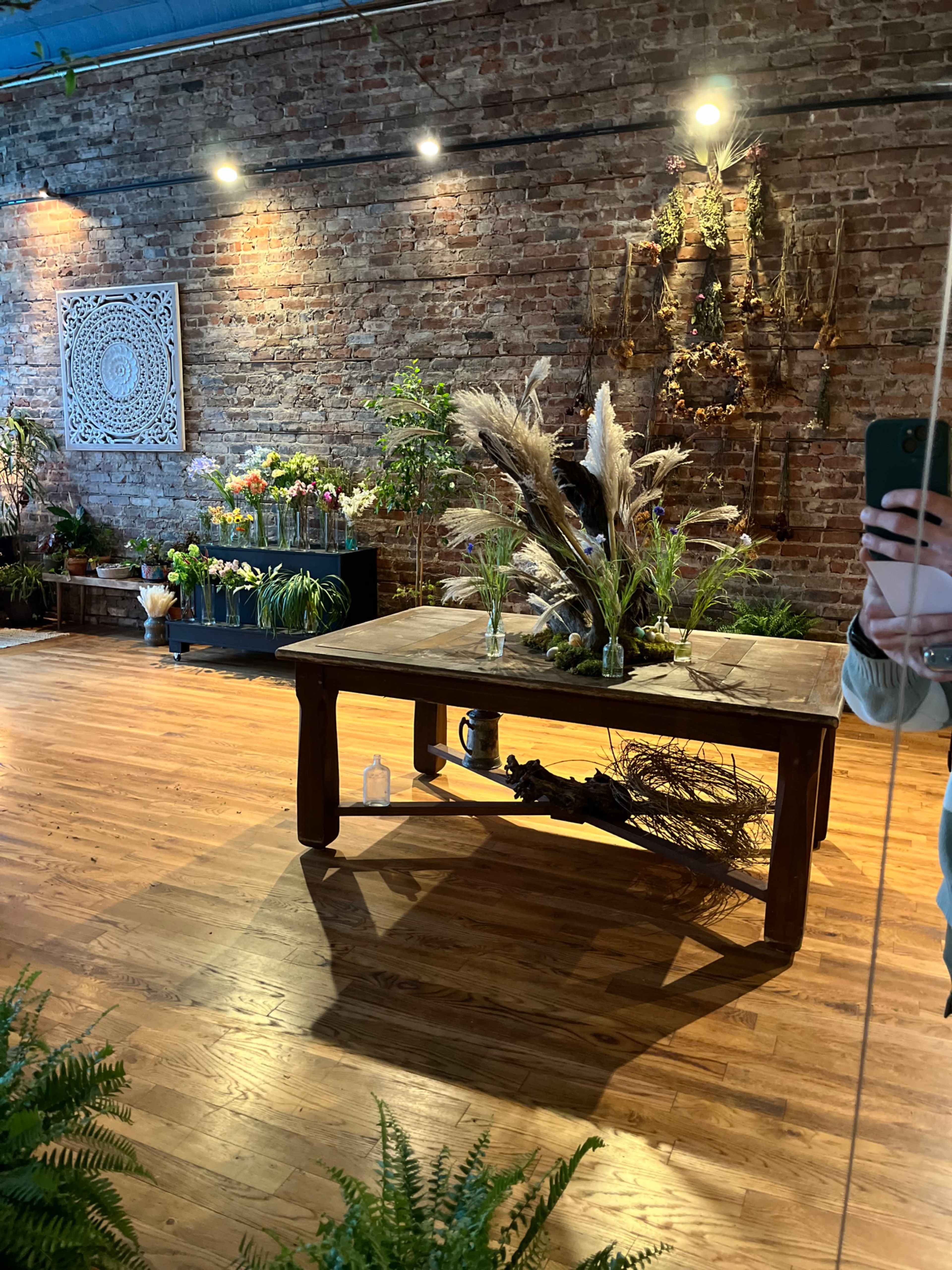 The image shows a floral shop interior with a wooden table at the center surrounded by various plants and flowers against a brick wall.