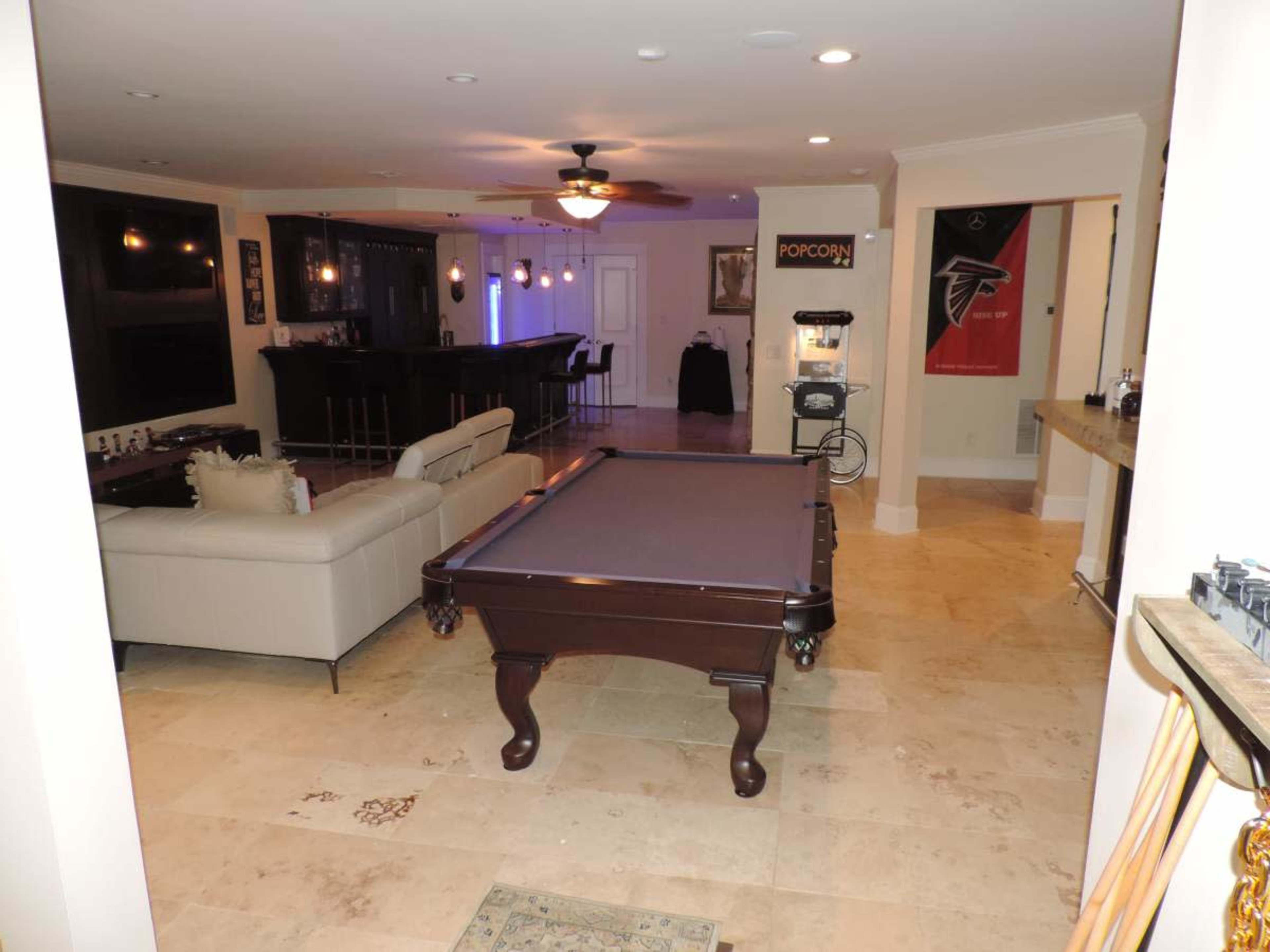 The image shows a spacious entertainment room featuring a pool table, a sectional sofa, a bar area, and a popcorn machine.