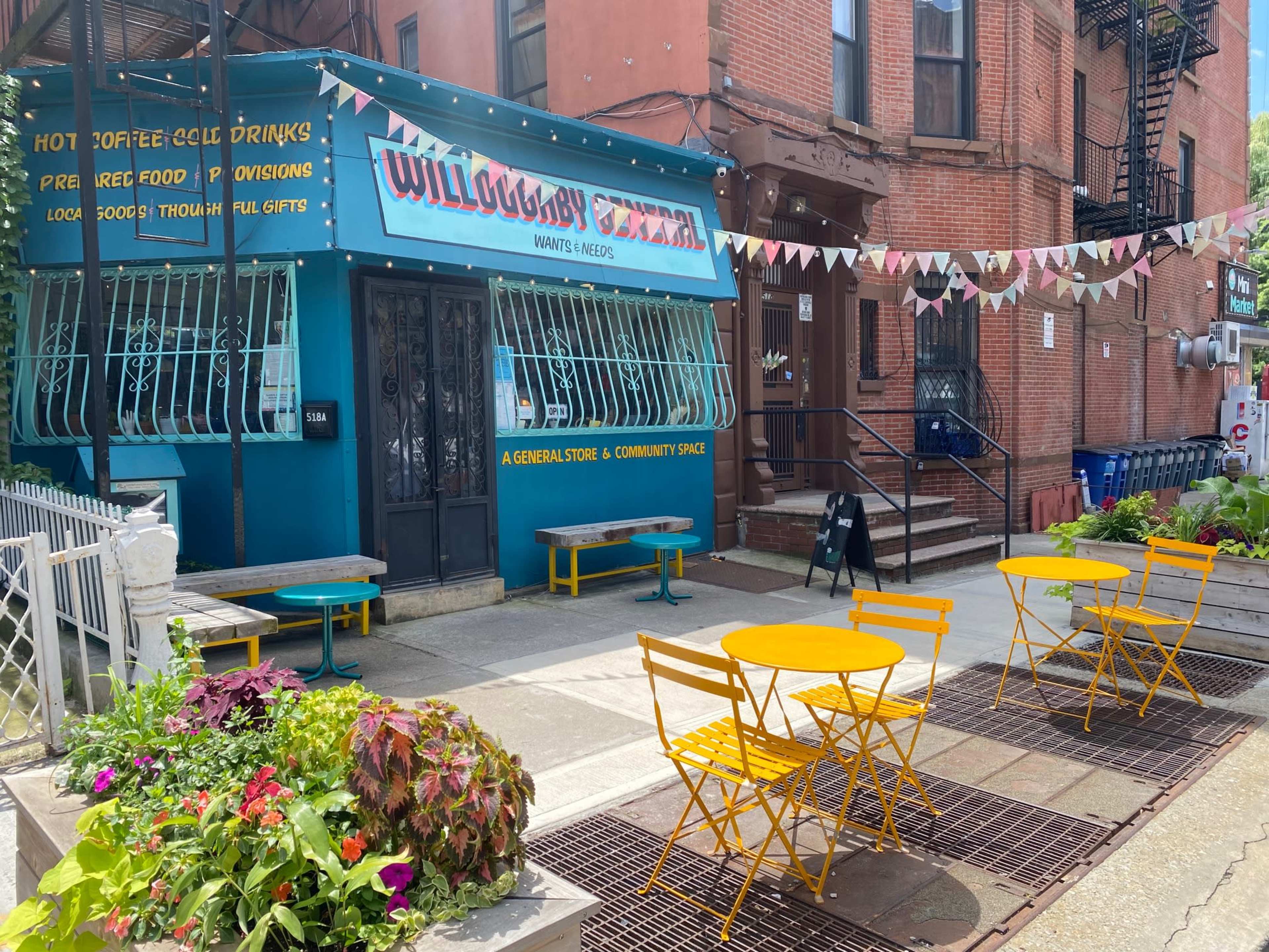 A colorful storefront with outdoor yellow tables and chairs, featuring a sign that reads "Willoughby General" and decorations hung above.